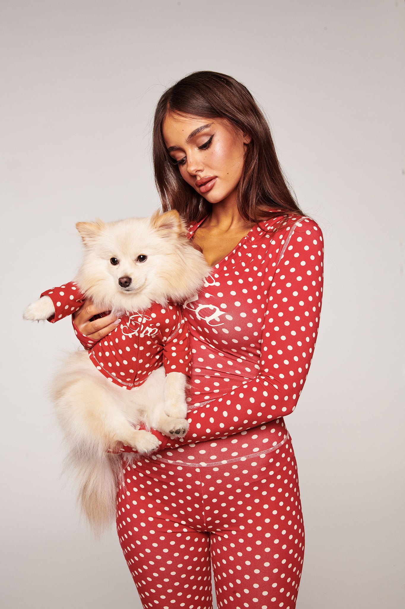 A woman wears red and white polka dot pajamas, holding her dog dressed in the I.AM.GIA BLARE BEST FRIEND DOG HOODIE - RED POLKA DOT. Both face the camera against a plain light background, showing their coordinated style.