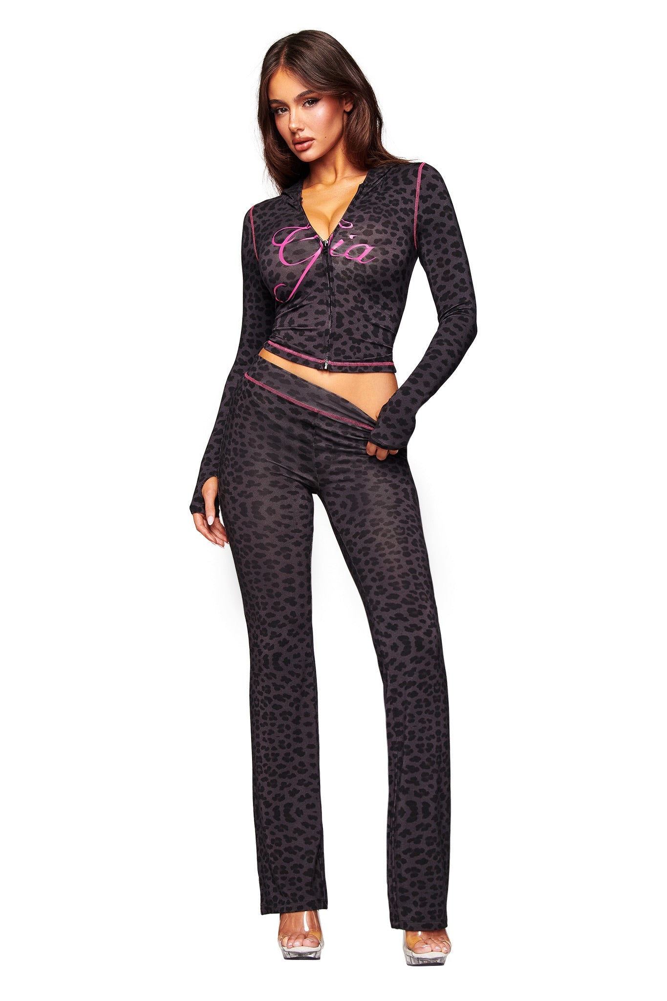 A woman wears the I.AM.GIA BLARE HOODIE - PANTHER, a fitted black leopard-print two-piece with long sleeves and flared pants. She pairs it with clear heels and poses against a white background.