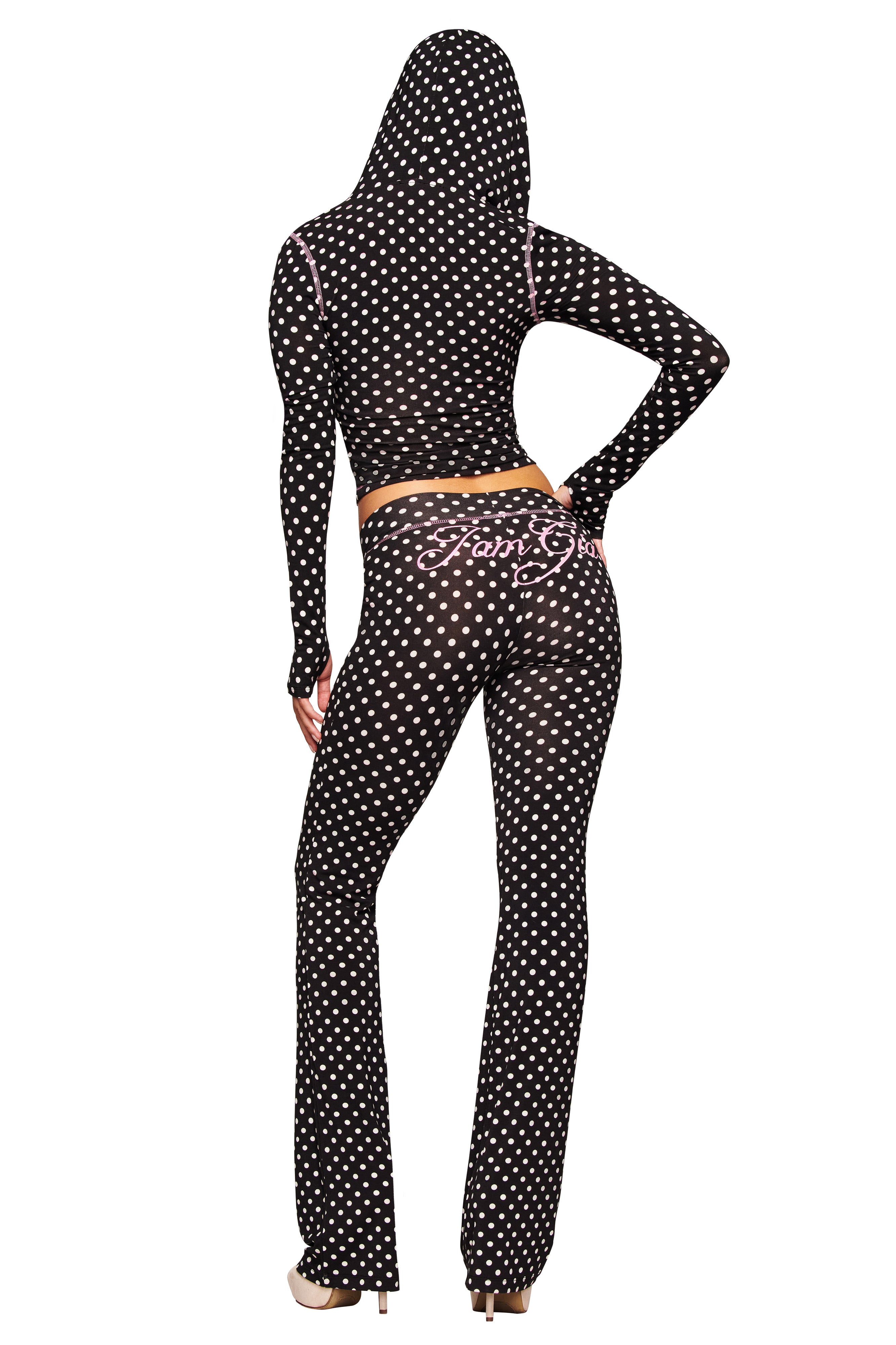 A person wearing I.AM.GIA's BLARE TRACKPANT - BLACK POLKA DOT stands against a white background, facing away with one hand on their hip, showcasing the black polka dot design and pink writing on the back.