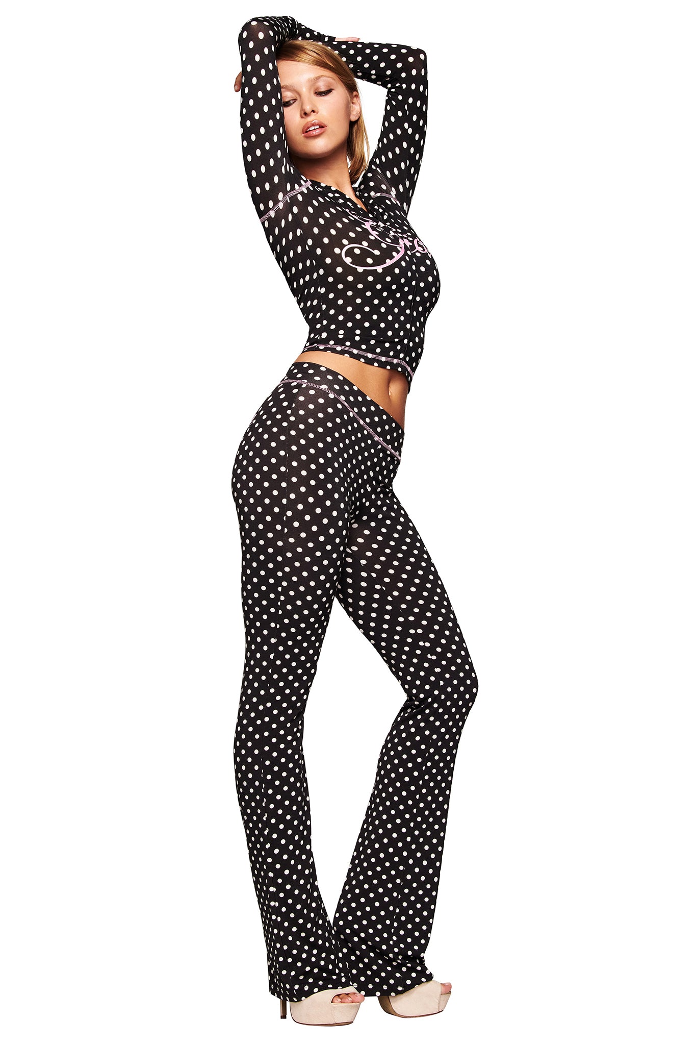 A woman models the I.AM.GIA BLARE HOODIE in black polka dot, paired with matching flared pants and high heels. She stands confidently in a side pose, one arm raised above her head.