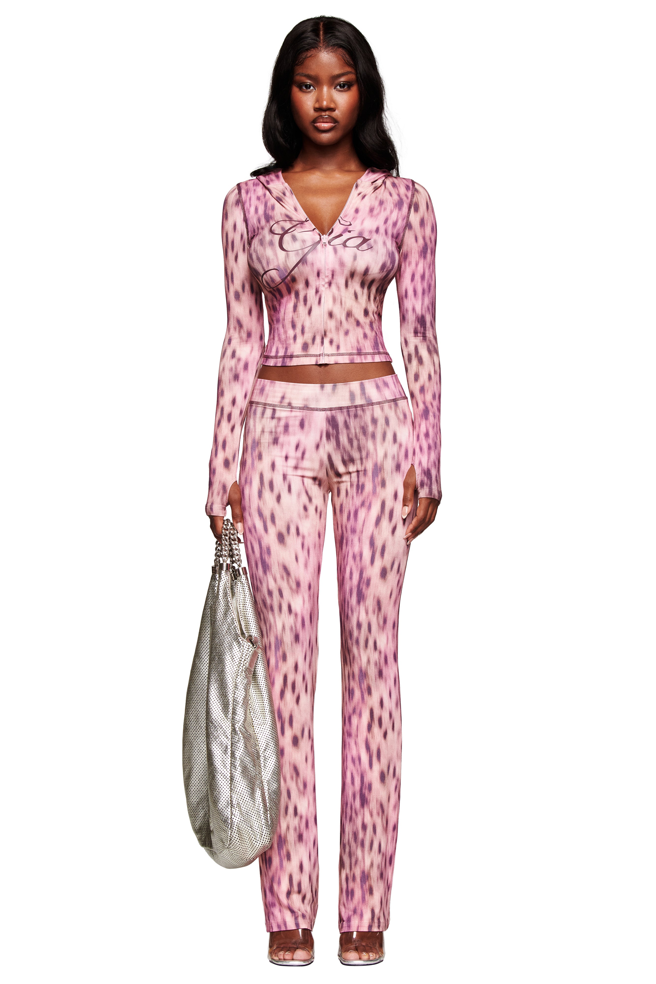 A woman stands facing forward against a white background, showcasing her bold style in the I.AM.GIA BLARE HOODIE - PINK BLURRED CHEETAH, paired with open-toe heels and holding a large silver textured bag.