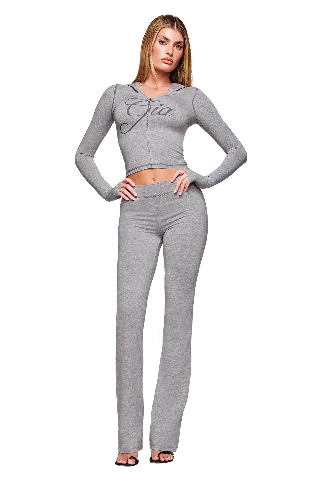 A woman wears I.AM.GIA's BLARE JERSEY TRACKPANT in heather grey, featuring a flared leg, paired with a matching cropped zip-up hoodie with "Gia" on the front. She has long straight hair and open-toe heels.