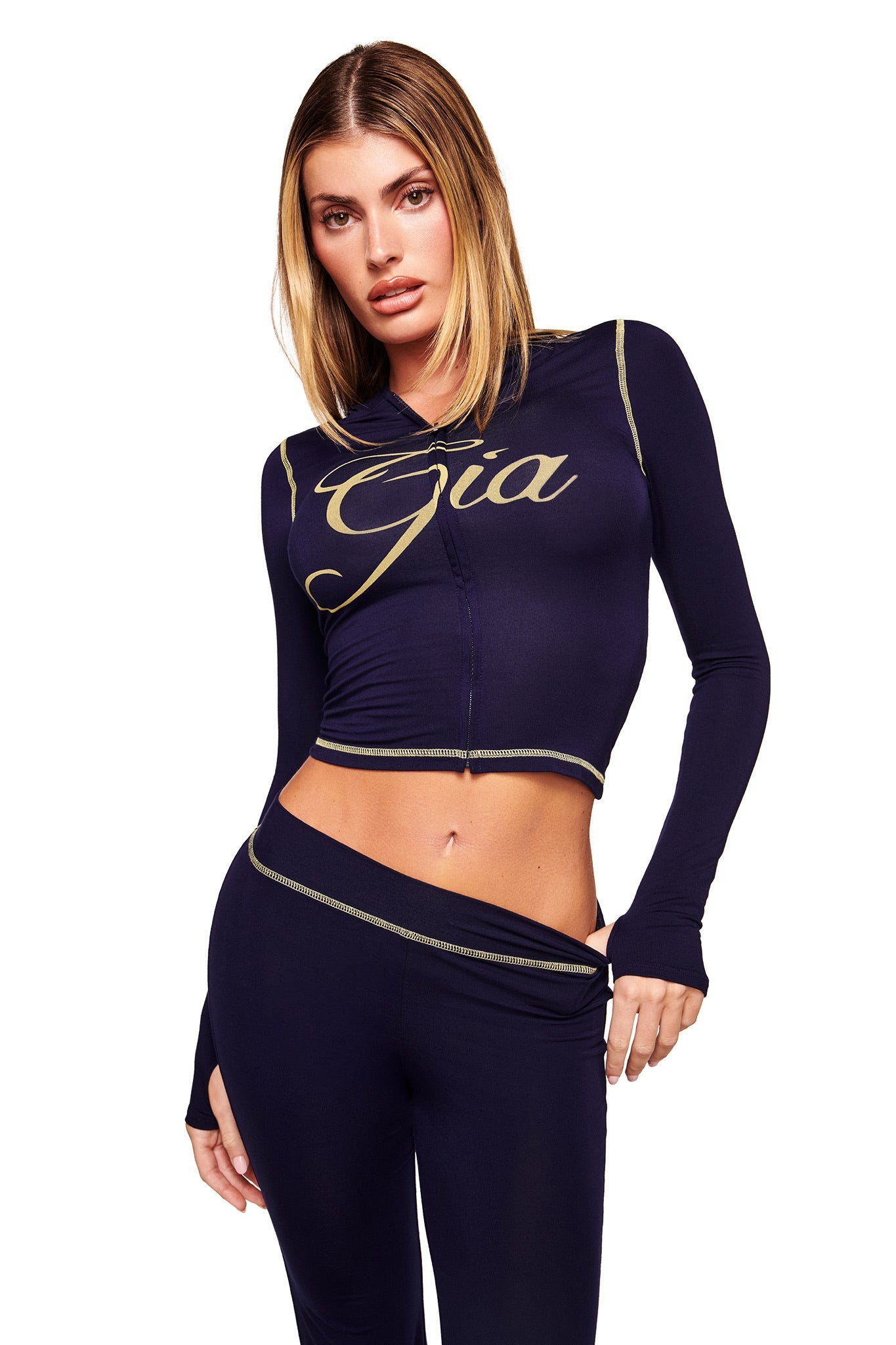 A woman wears the BLARE HOODIE in navy by I.AM.GIA, featuring gold stitching and "Gia" in bold gold script across the chest. She stands against a white background.