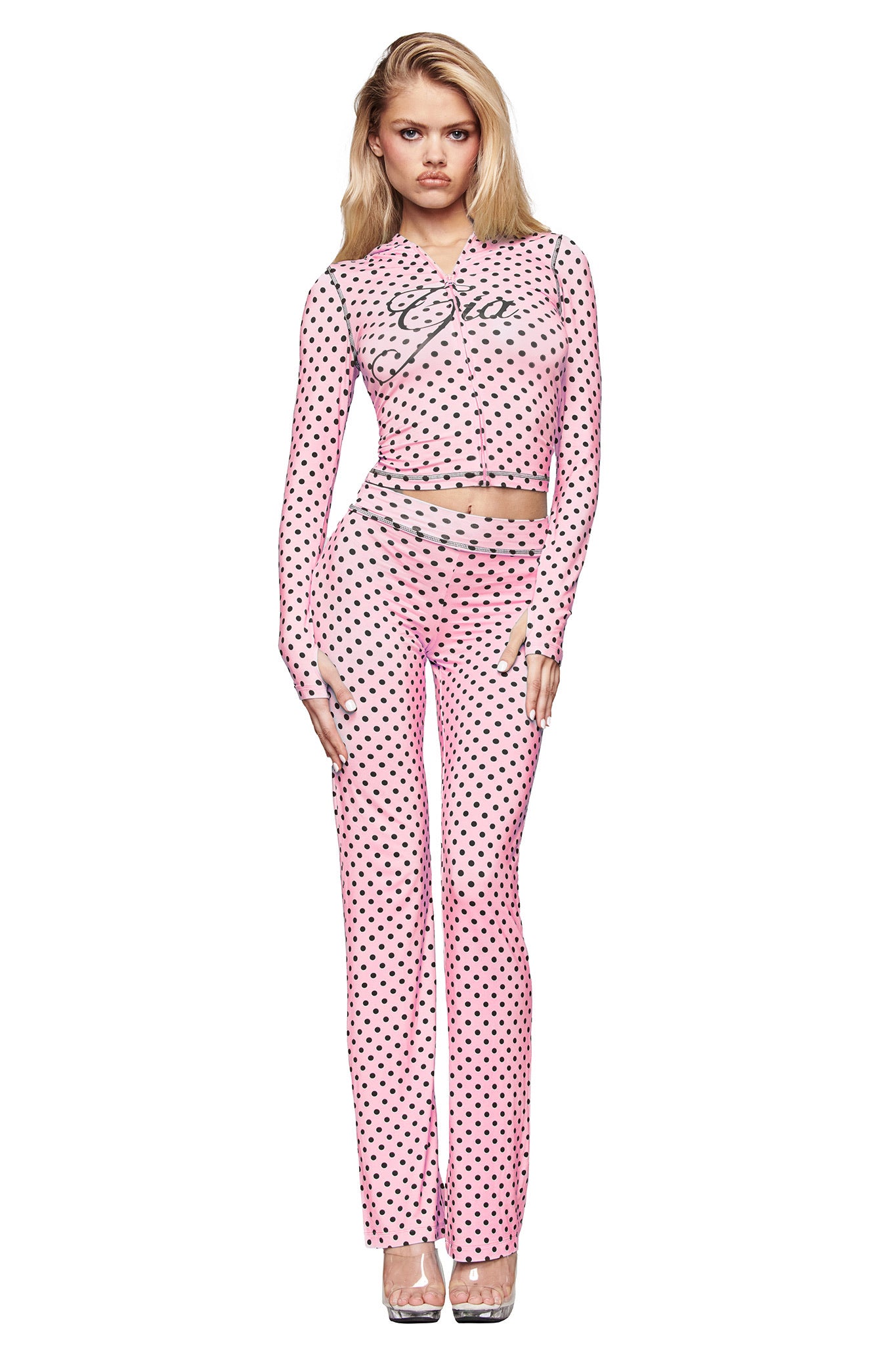 A woman with long blonde hair faces forward in a fitted, long-sleeve pink top and matching I.AM.GIA BLARE TRACKPANT - PINK POLKA DOT. She wears clear heels against a white background, with the I.AM.GIA logo subtly accenting her look.