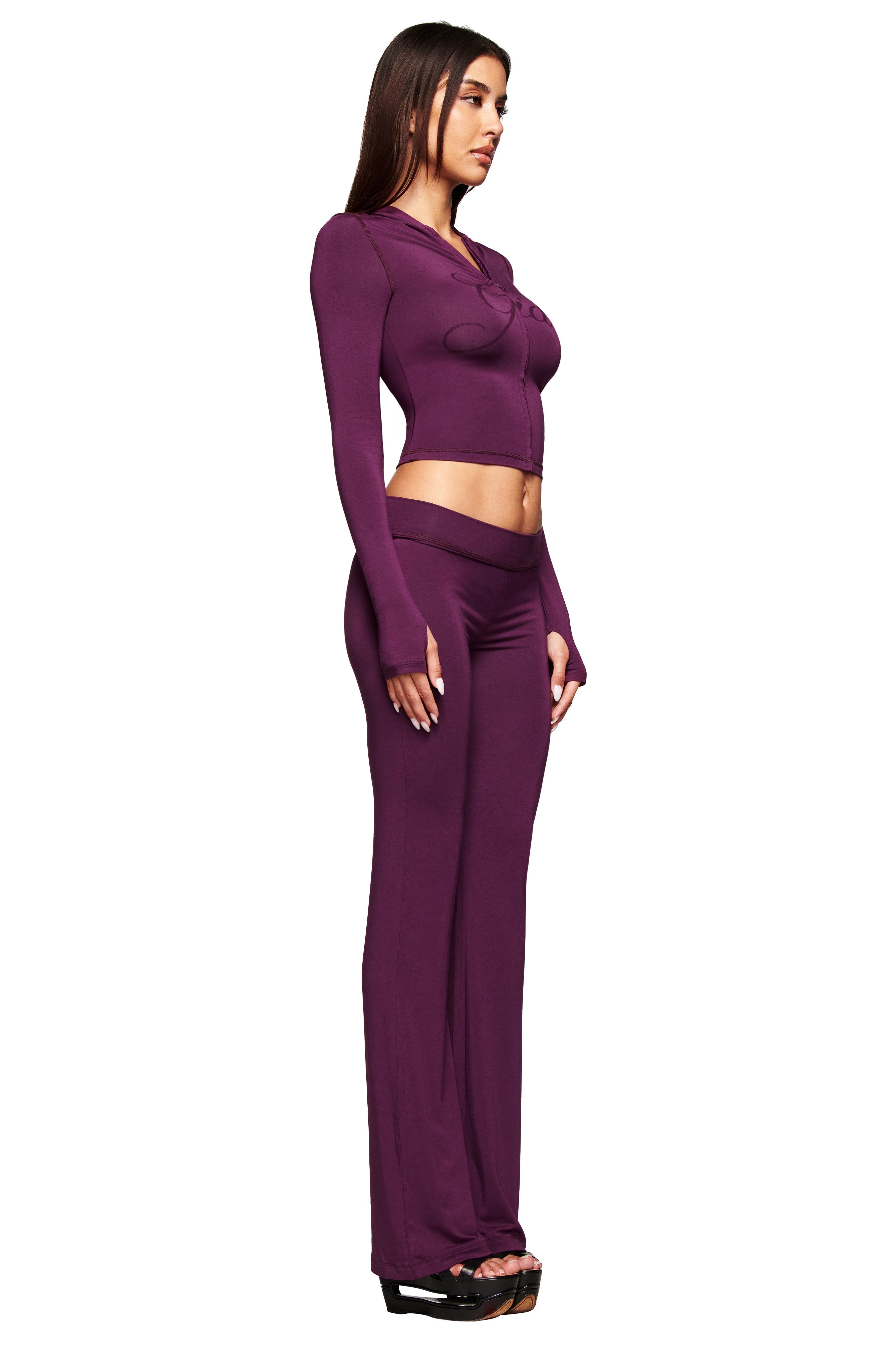 A woman stands in profile wearing the I.AM.GIA BLARE TRACKPANT in plum, styled with a fitted purple crop top and black open-toe heels, against a white background.