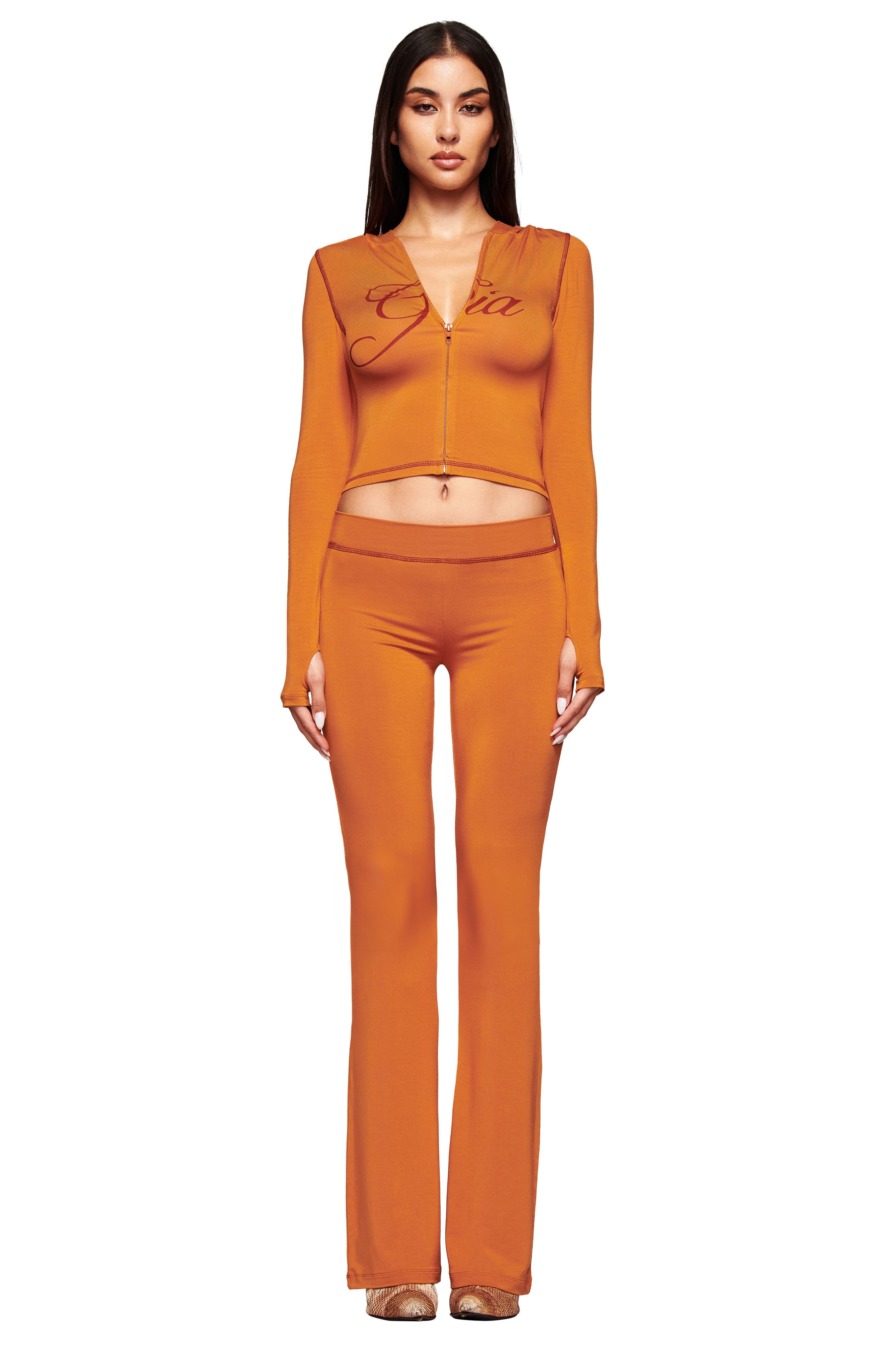A woman faces forward in the I.AM.GIA BLARE HOODIE - PUMPKIN set, featuring a long-sleeve orange zip-up crop top with "Gia" on the front and matching flared pants. She wears her straight brown hair down and has a neutral expression.