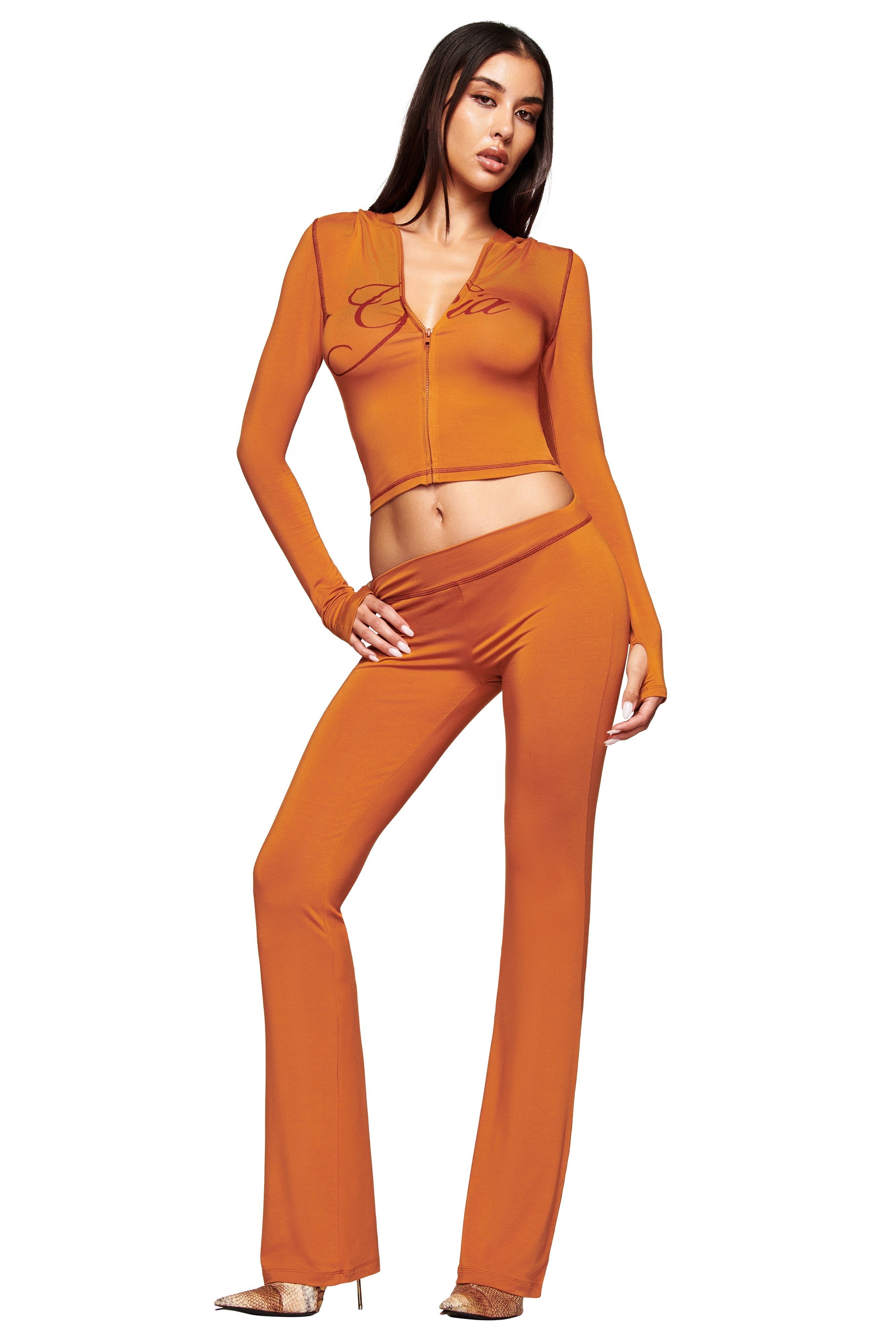 A woman poses confidently in the I.AM.GIA BLARE HOODIE - PUMPKIN, an orange zip-up crop top with matching high-waisted flared pants and beige heels, against a white background.