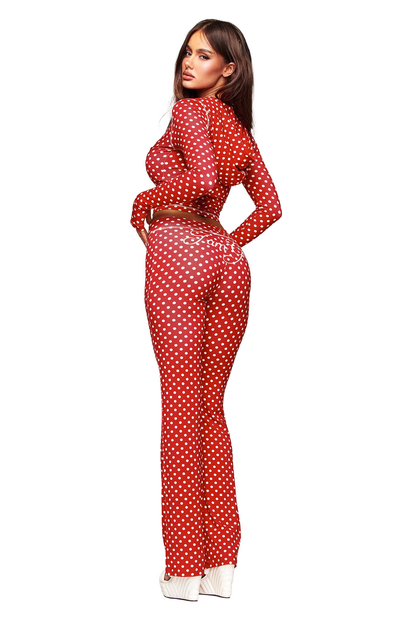 A woman poses against a white background wearing the I.AM.GIA BLARE HOODIE - RED POLKA DOT, a long-sleeved fitted jumpsuit, styled with white platform shoes and glancing over her shoulder.