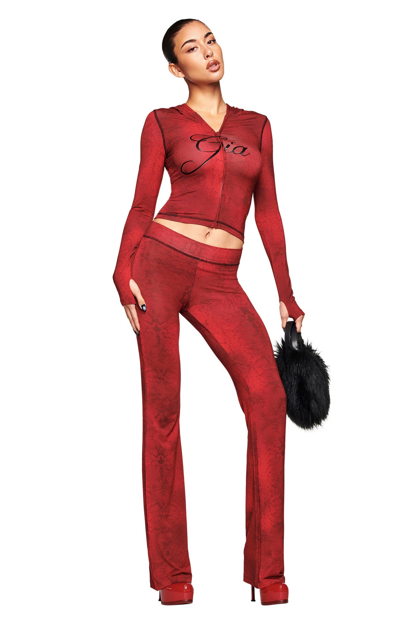 A woman models the I.AM.GIA BLARE HOODIE in red serpentine, a fitted two-piece set with long sleeves and flared pants. She pairs it with red platform heels, a black furry handbag, and a top featuring "Gia" in black script on white.