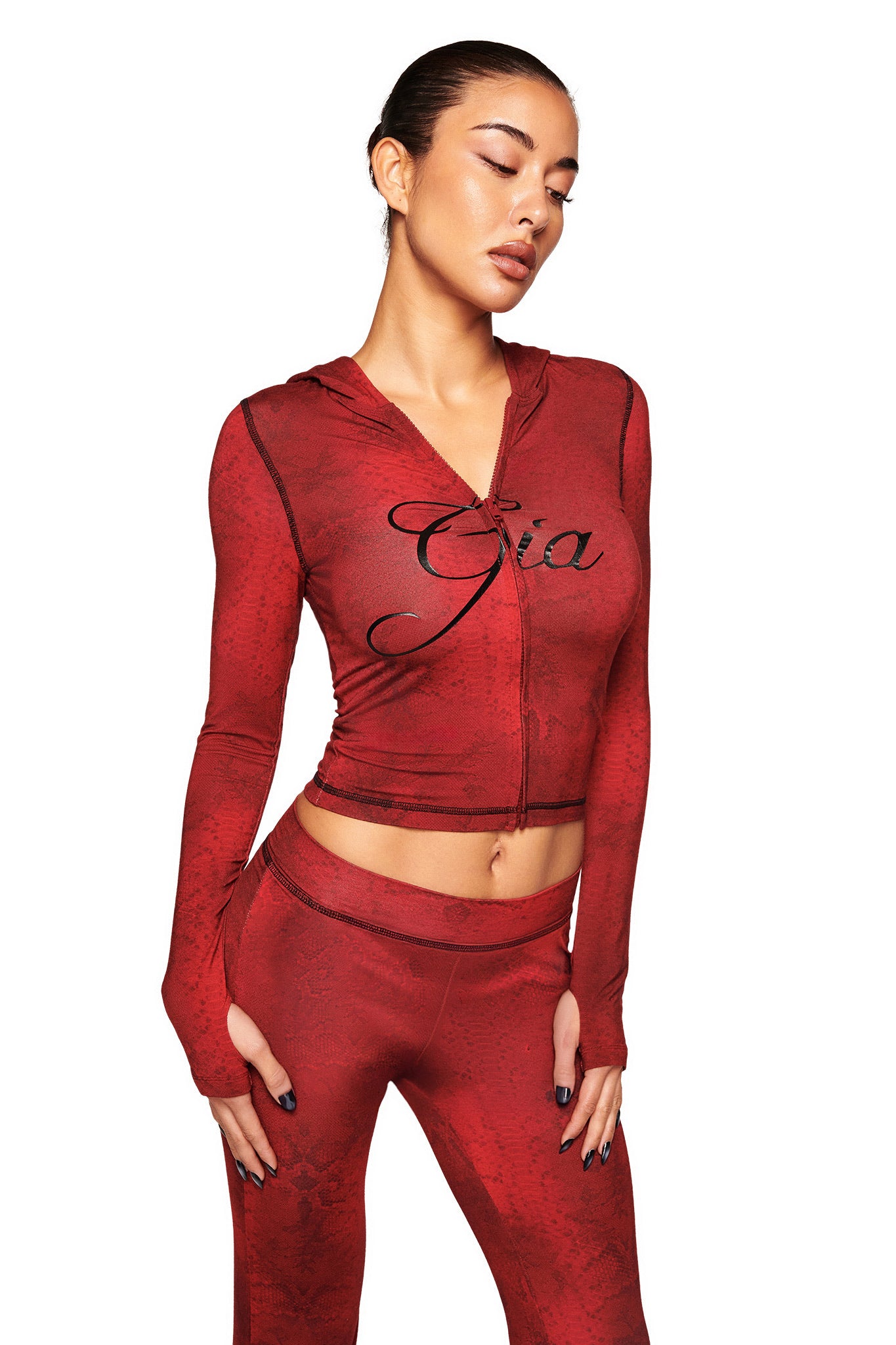 A woman in a fitted I.AM.GIA BLARE HOODIE – RED SERPENTINE with matching red serpentine print pants, "Gia" on the chest, stands against a white background with her head tilted and eyes closed.