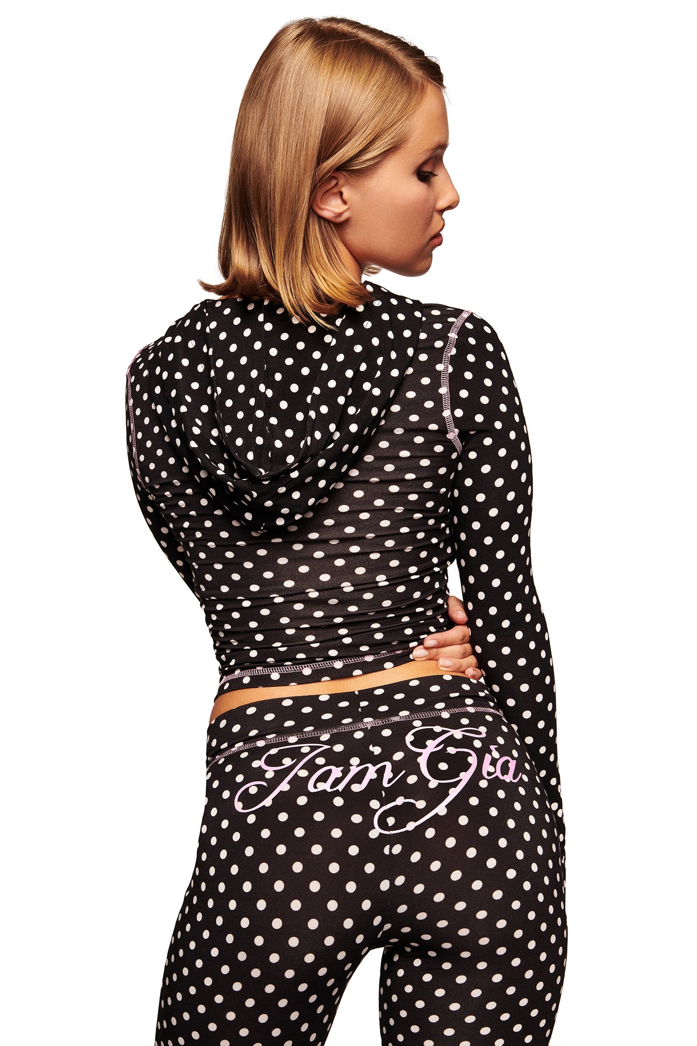 A person with short blonde hair stands turned to the side, wearing a black long-sleeved hooded top and I.AM.GIA's BLARE TRACKPANT in black polka dot with white polka dots and cursive writing across the back.