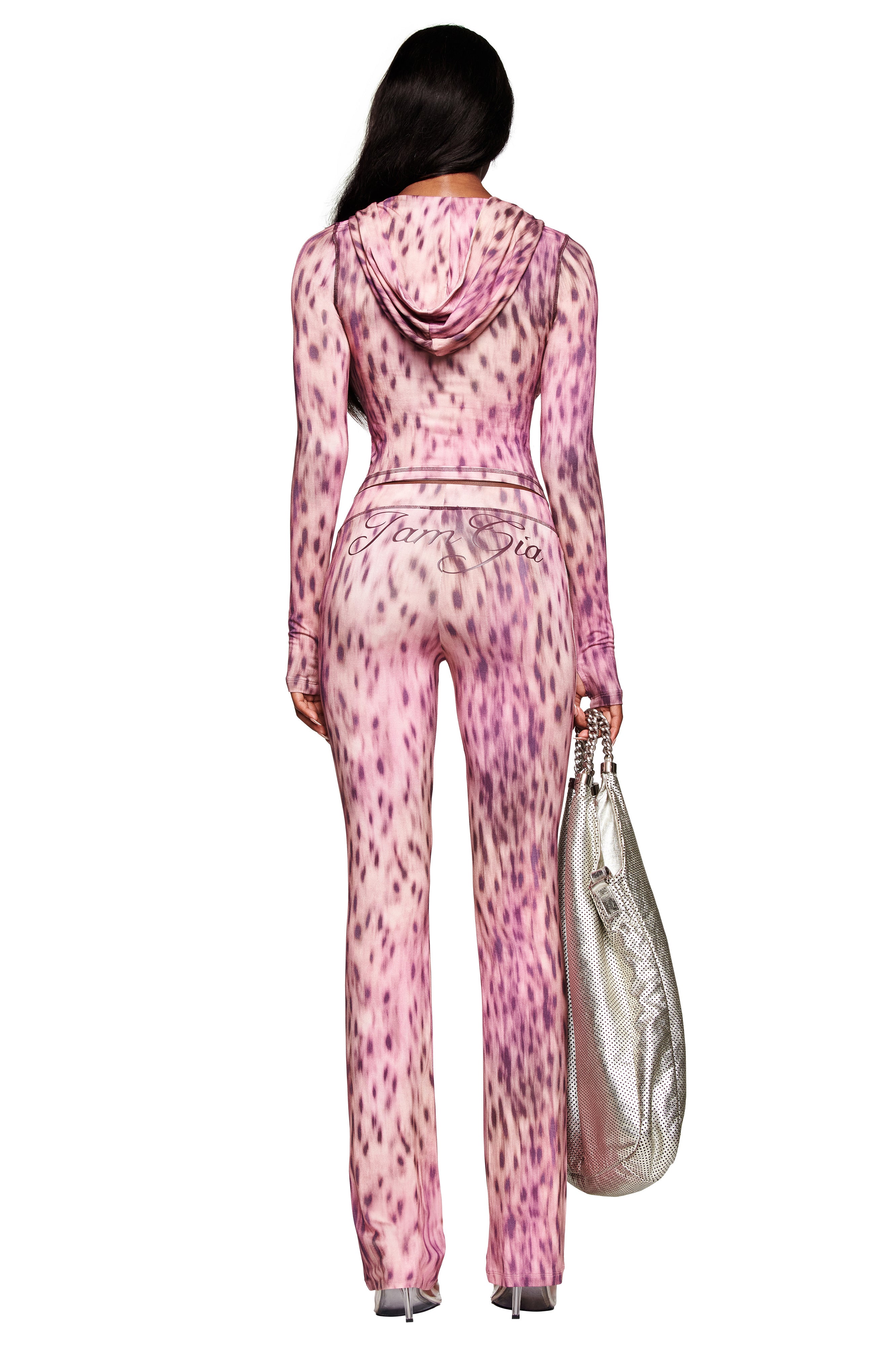 A woman stands with her back to the camera, wearing I.AM.GIA’s BLARE TRACKPANT in pink blurred cheetah print and holding a large silver textured bag. The outfit features "ArteSia" written in script on the back.