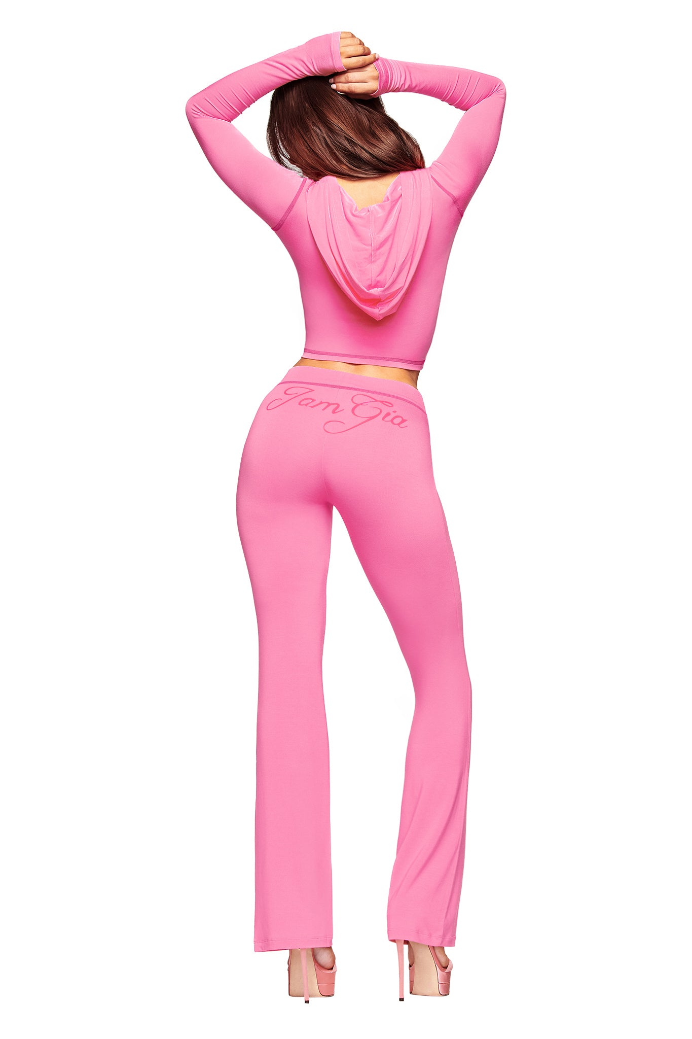 A woman faces away from the camera, modeling I.AM.GIA’s BLARE HOODIE in neon pink—a bodycon crop top with matching flared pants featuring "Pam Gia" on the back—posing in high heels, arms elegantly raised overhead.