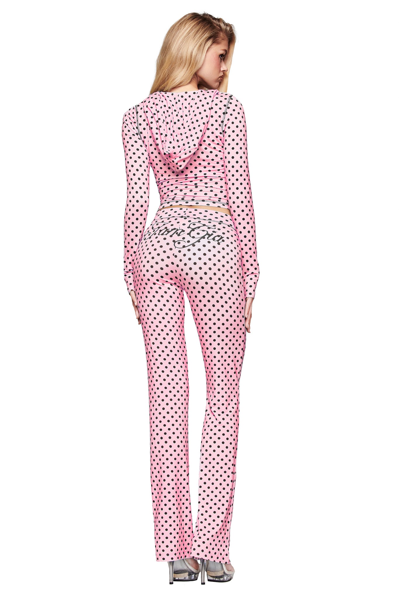 A woman faces away in the I.AM.GIA BLARE TRACKPANT - PINK POLKA DOT and a sheer pink hooded top, both featuring silver script logo details. She finishes the look with silver heels against a white background.