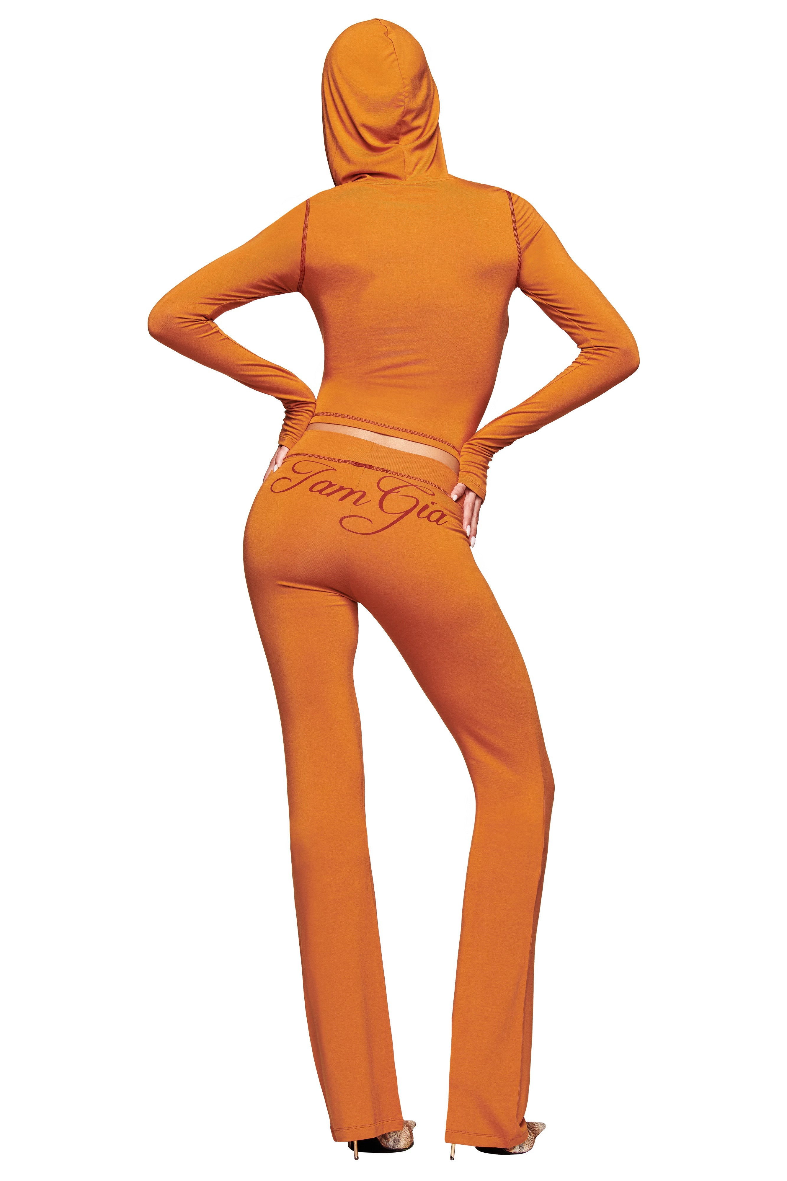 A person stands hands-on-hips, back to the camera, wearing the I.AM.GIA BLARE HOODIE - PUMPKIN and matching high-waisted pants with "Farm Gia" on the back, set against a white background.