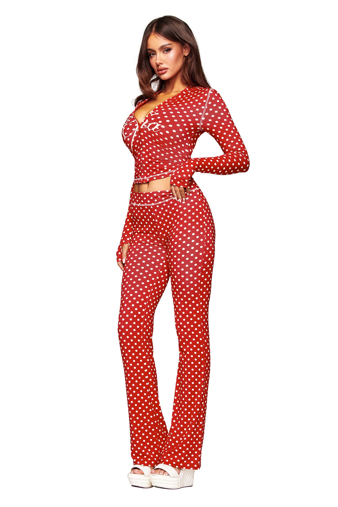 A woman poses against a white background wearing the I.AM.GIA BLARE TRACKPANT in red polka dot, styled with a matching crop top, long brown hair, and white platform shoes.