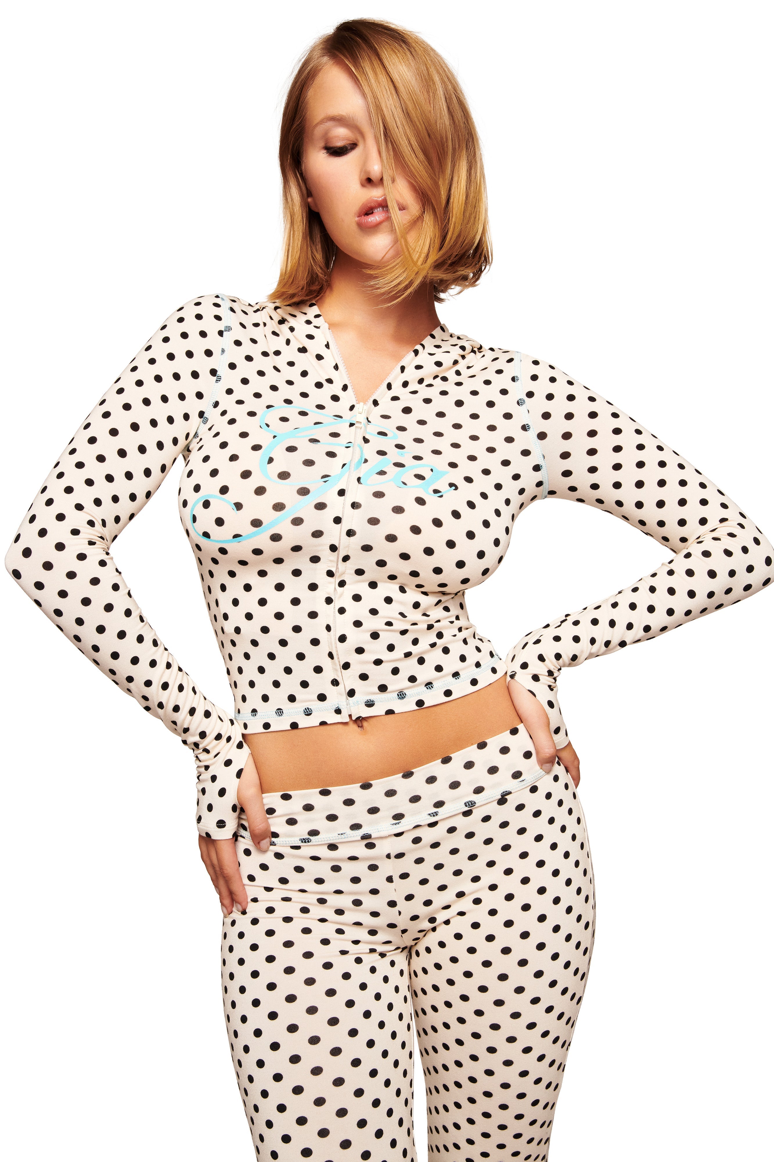 A woman with shoulder-length blonde hair poses with one hand on her hip, wearing the I.AM.GIA BLARE TRACKPANT in cream polka dot, a fitted long-sleeve white outfit with flared legs, set against a white background.