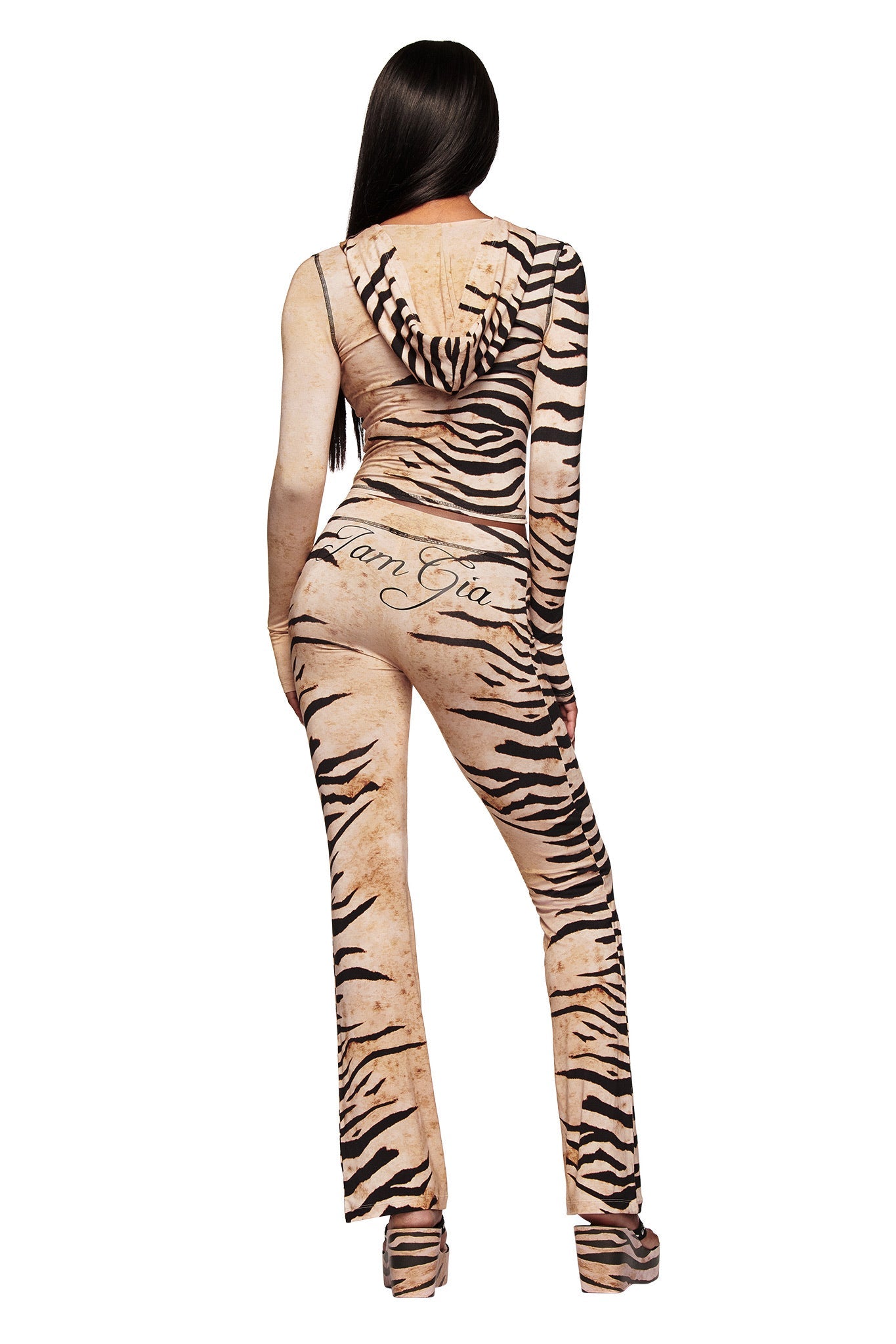 A woman with long dark hair stands facing away, wearing the BLARE HOODIE - WILD SIDE jumpsuit from I.AM.GIA and platform heels. The beige tiger-striped bamboo piece features "I Am Gia" on the back.