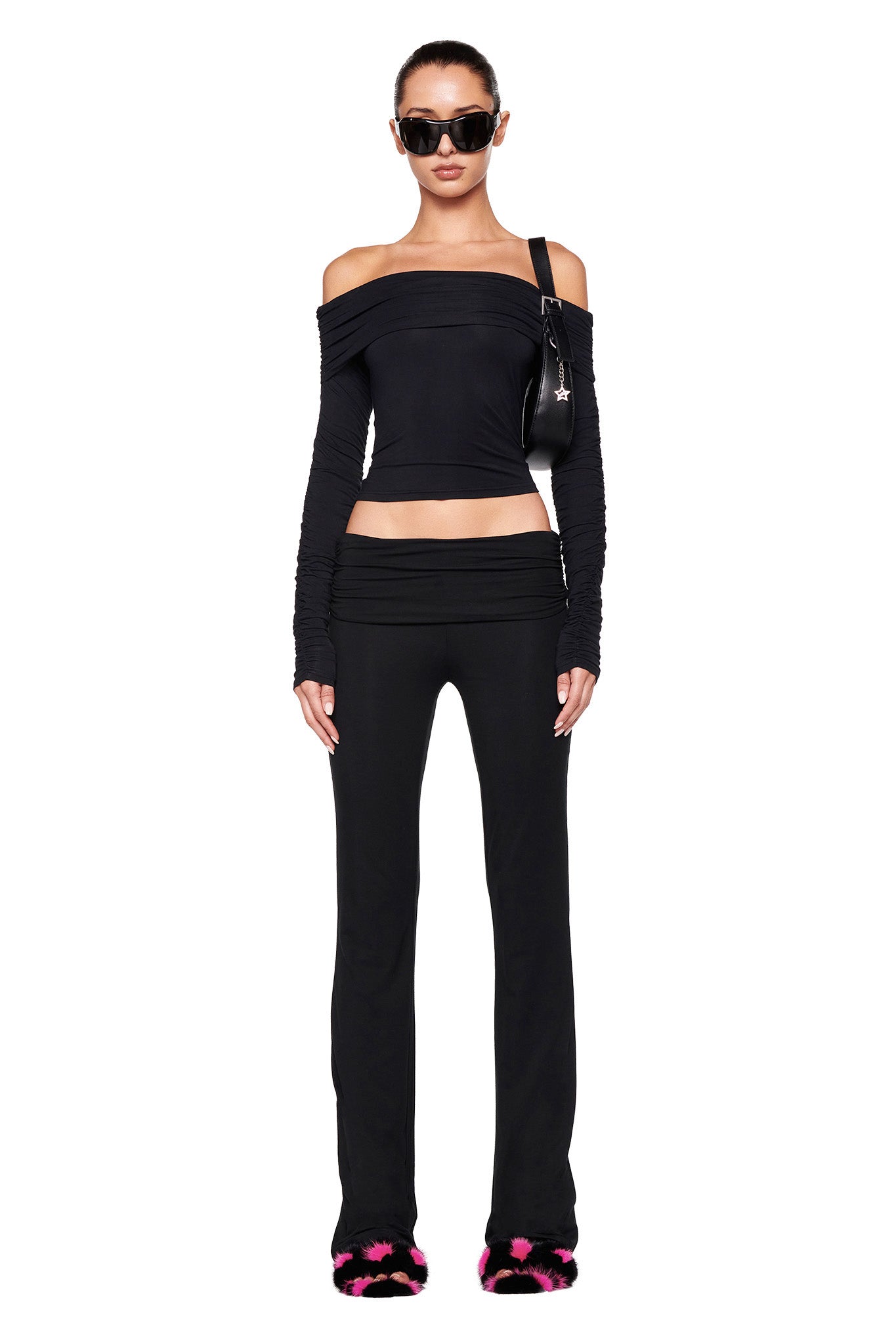 A person stands against a white background wearing an I.AM.GIA off-shoulder black crop top with long sleeves, matching I.AM.GIA JANNIS PANT in solid black, black sunglasses, and black shoes with pink accents. The person holds a black shoulder bag and their hair is pulled back.