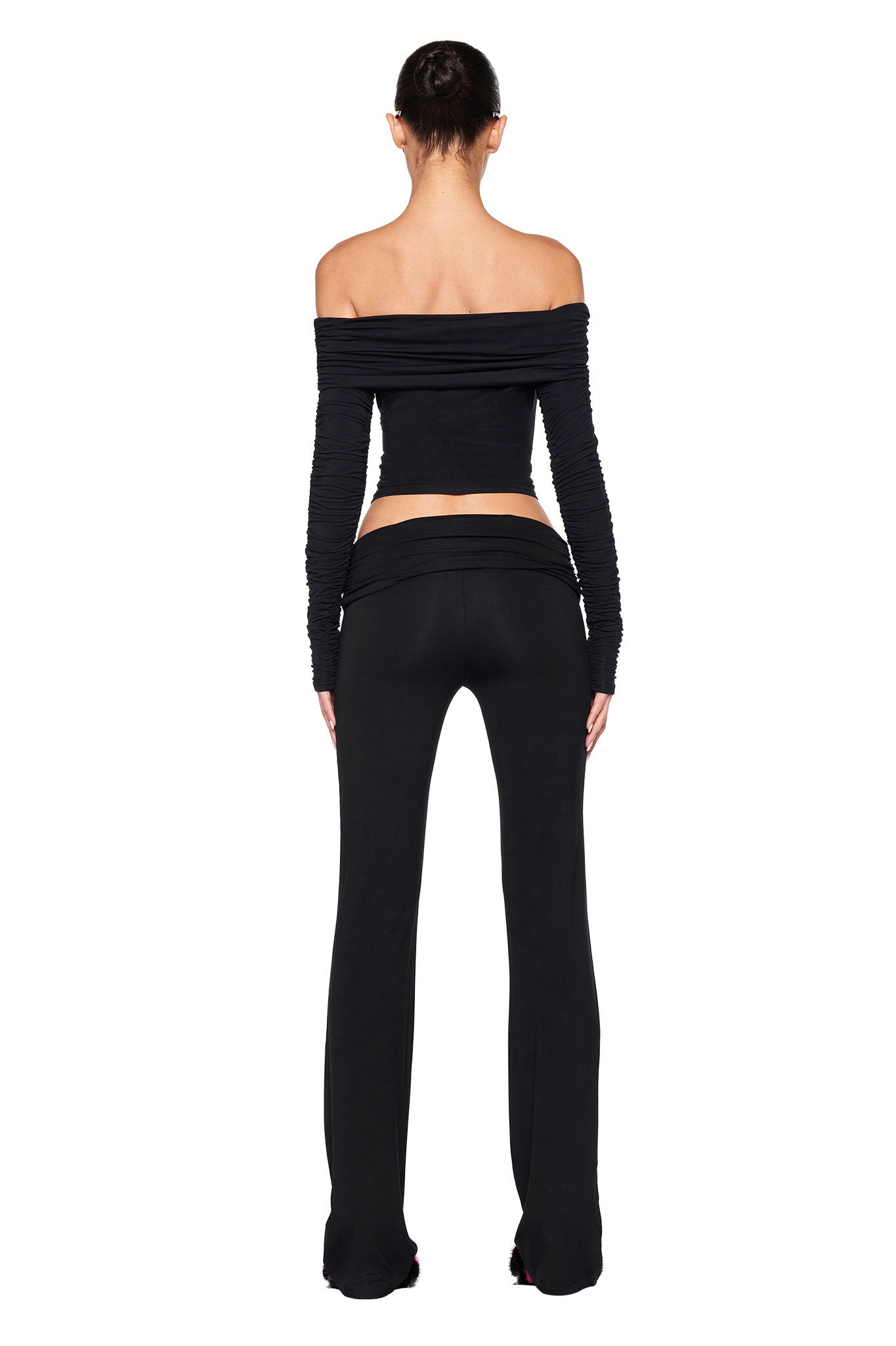 A person is standing with their back to the camera, wearing a black off-the-shoulder long-sleeve top and the JANNIS PANT - BLACK by I.AM.GIA. Their hair is tied up in a bun, and the outfit features a cutout design at the back, revealing some skin around the waist area.
