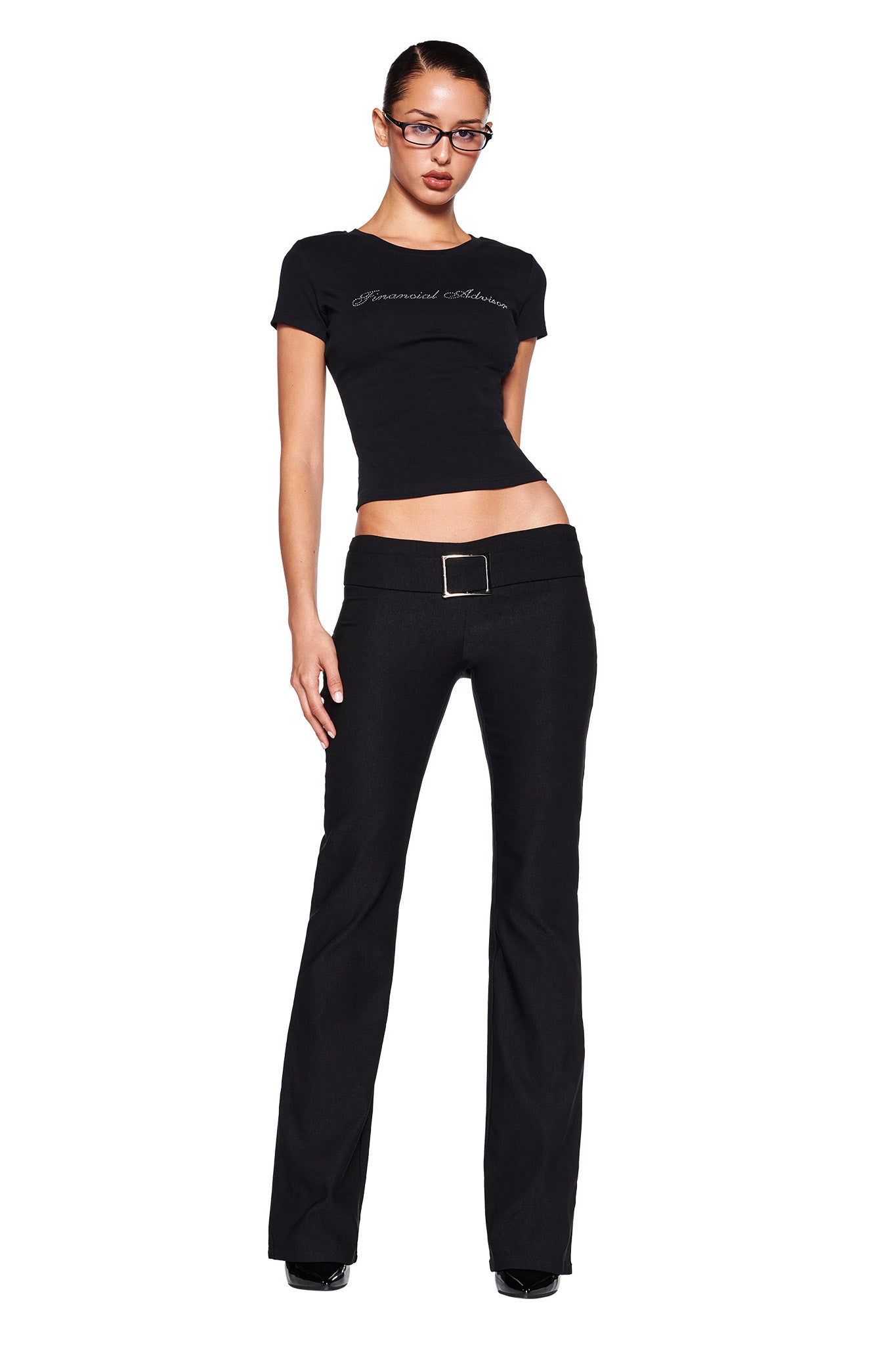 A person is standing against a white background, wearing a fitted black T-shirt and I.AM.GIA's SHARAH PANT - BLACK, featuring a low-rise waistline and a belt. They have glasses and are looking directly at the camera with a confident pose.