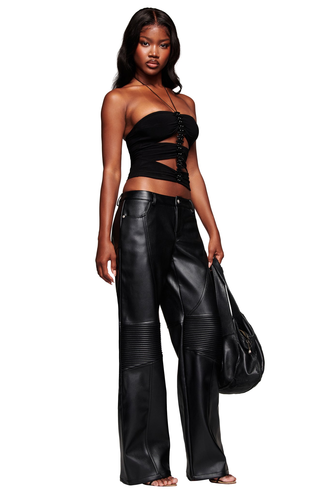 A woman with loose, wavy hair and a confident expression poses against a white background wearing I.AM.GIA's KRISTIE PANT in black—faux leather, wide-leg, low-rise style—paired with a strapless black cutout top and holding a black handbag.