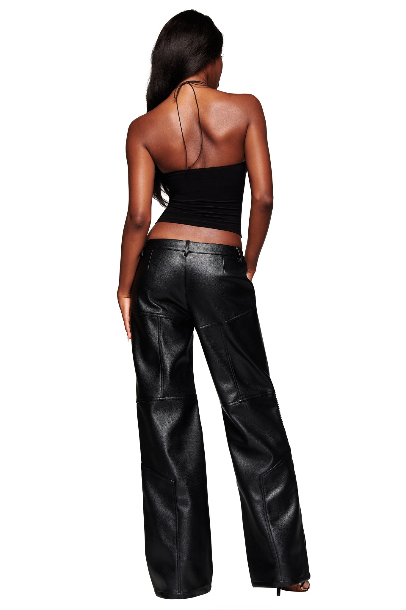 A woman with long dark hair faces away, wearing the I.AM.GIA KRISTIE PANT in black—a low-rise, faux leather style with seam details—paired with a black halter top against a white background.