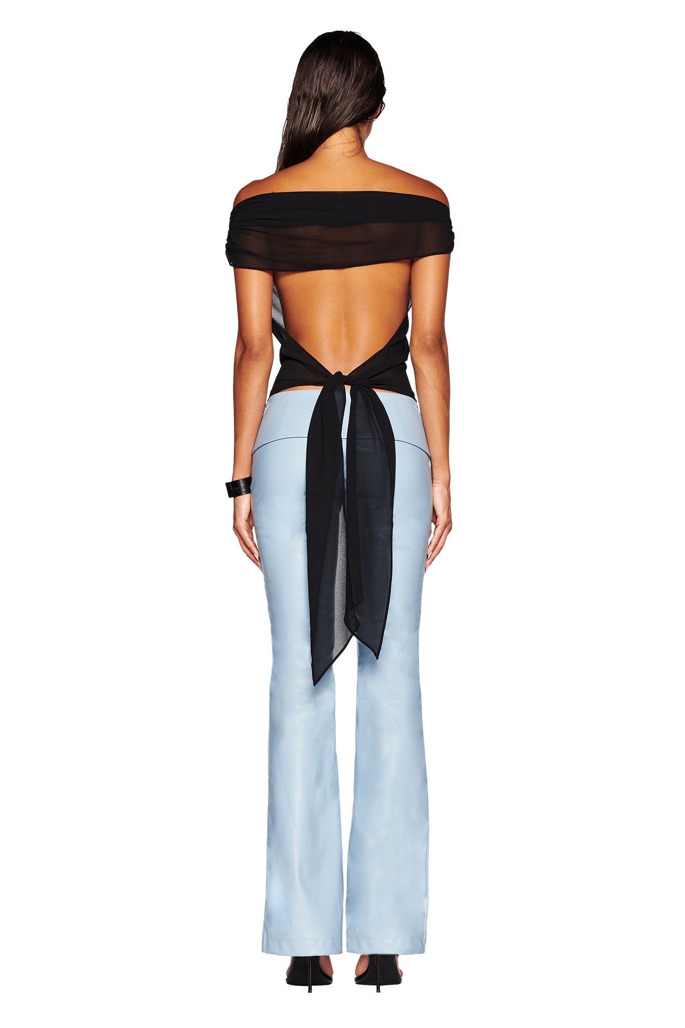 A person with long dark hair is shown from the back wearing the KAHLO TOP - BLACK by I.AM.GIA, an off-shoulder top made of georgette fabric, featuring an open back and a knot tied at the waist. This is paired with light blue high-waisted flare pants in a relaxed fit and black heeled shoes.