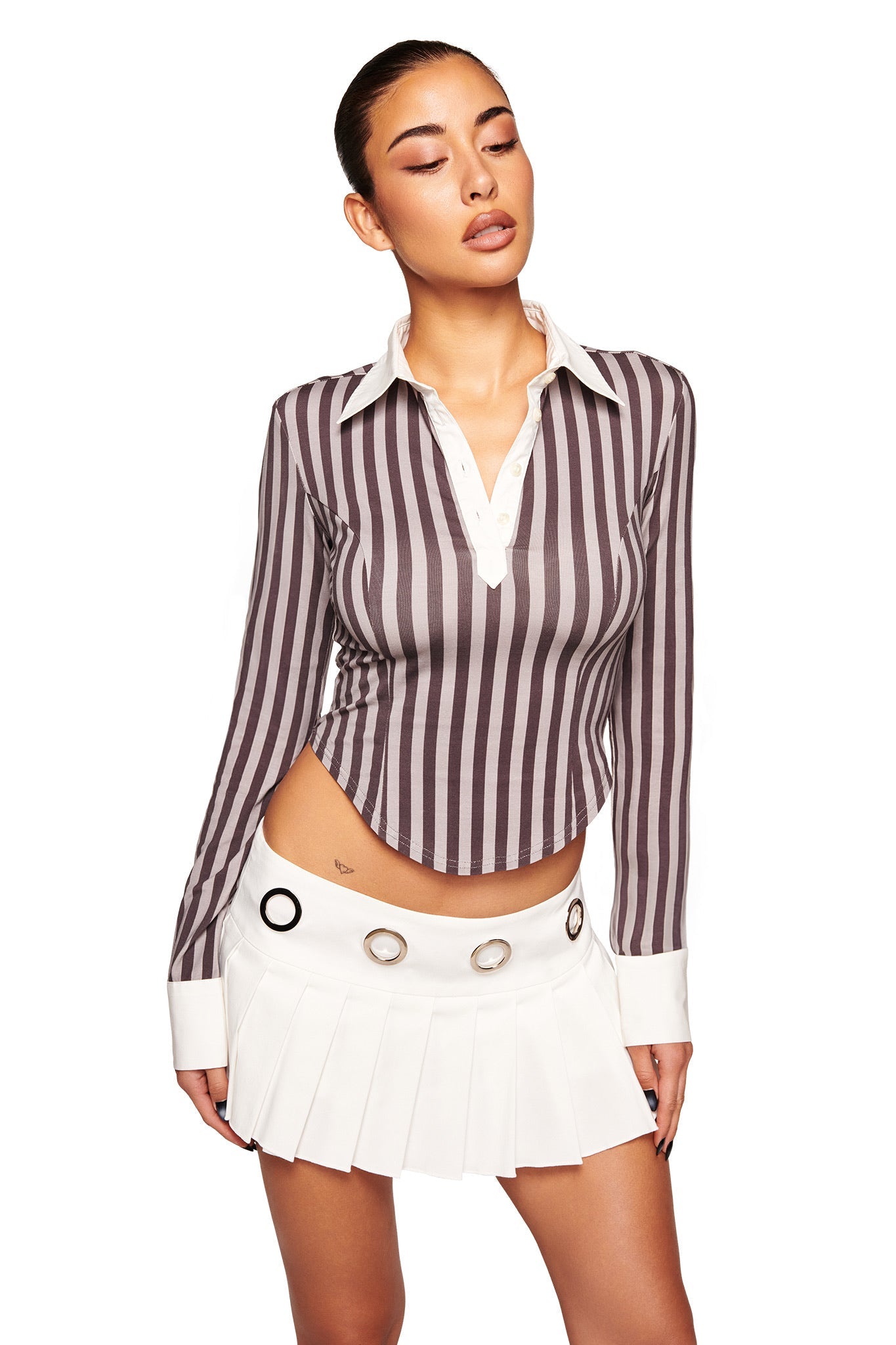 A woman poses in the I.AM.GIA JAYMES SKORT - WHITE, a short, white pleated skort with large metallic grommets and a trendy low-rise waist, paired with a long-sleeve brown and white striped blouse against a white background.