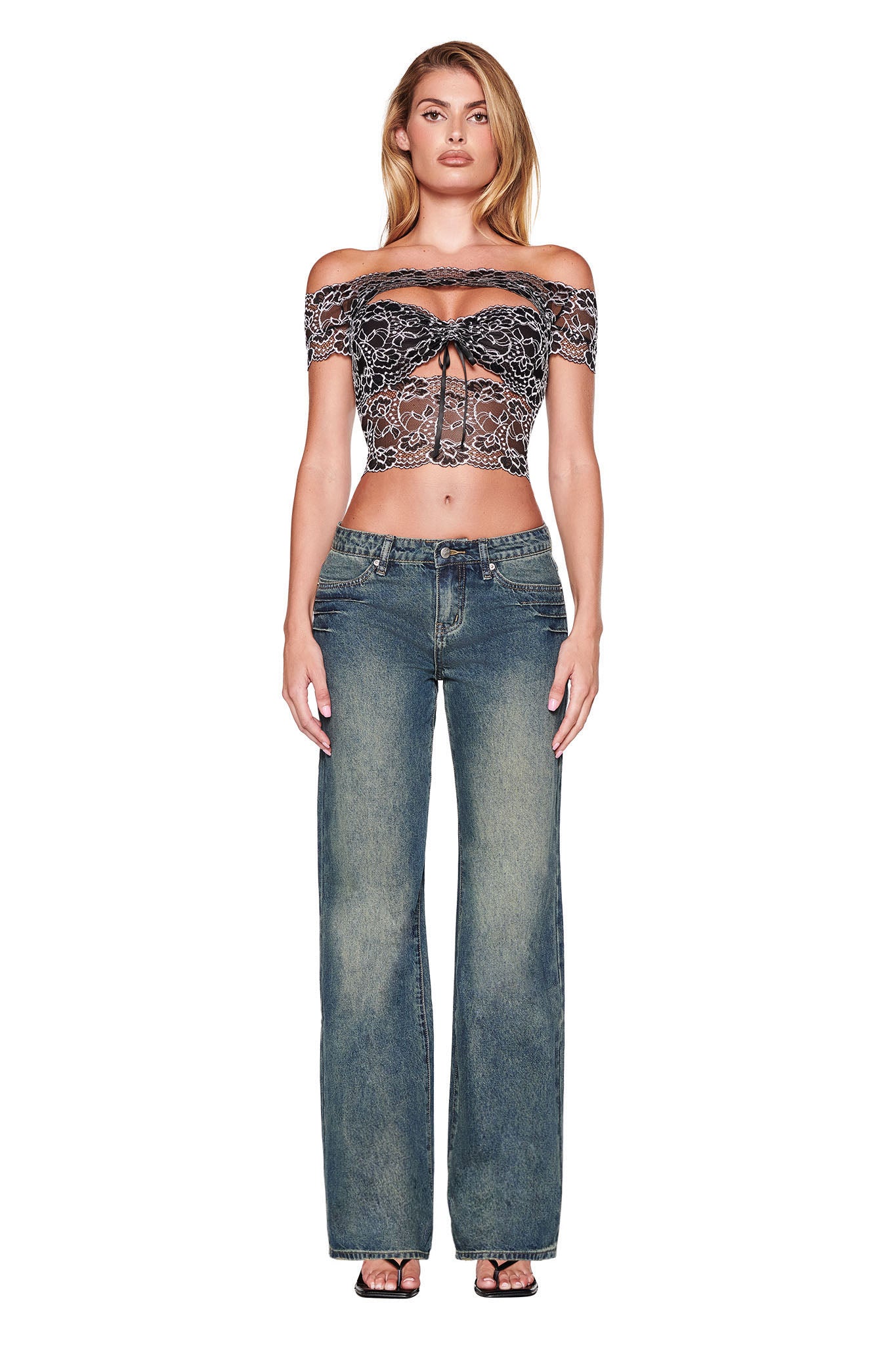 A person stands against a white background wearing an off-the-shoulder black lace top and the DYLAN JEAN in blue denim by I.AM.GIA. They have long hair and are wearing strappy black sandals.