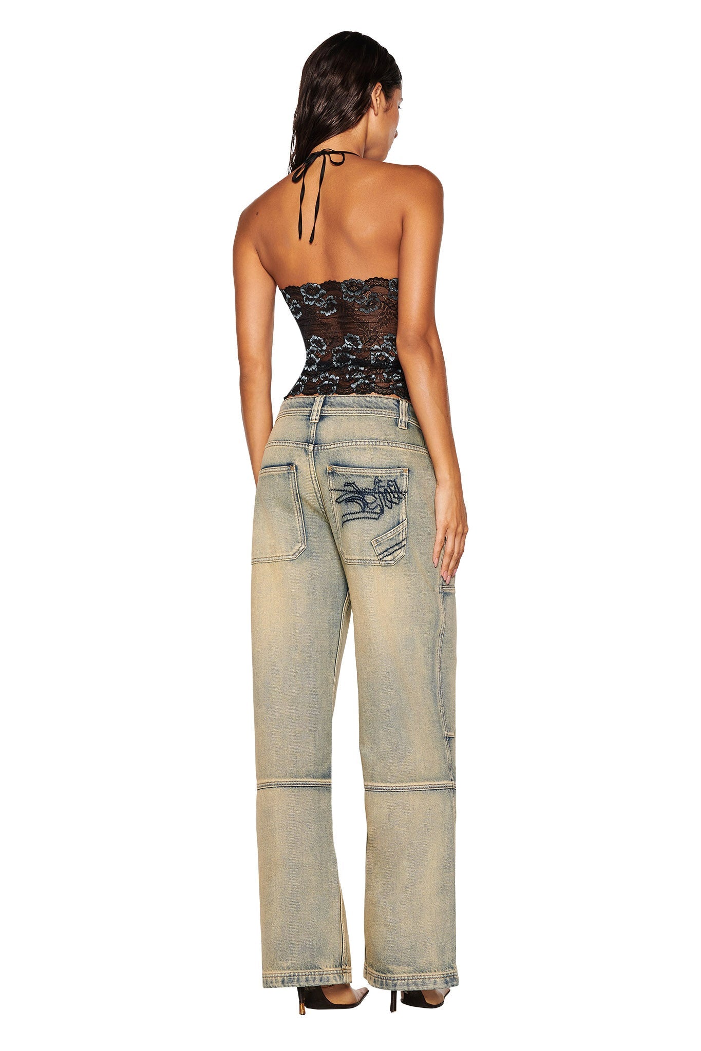 An individual is facing away wearing the GEM HALTER TOP - BLACK by I.AM.GIA, a black lace fabrication halter top. They have paired this with light blue wide-leg jeans that feature distressed patches and embroidery. Their long hair flows down their back, and they are in high-heeled shoes against a plain white backdrop.