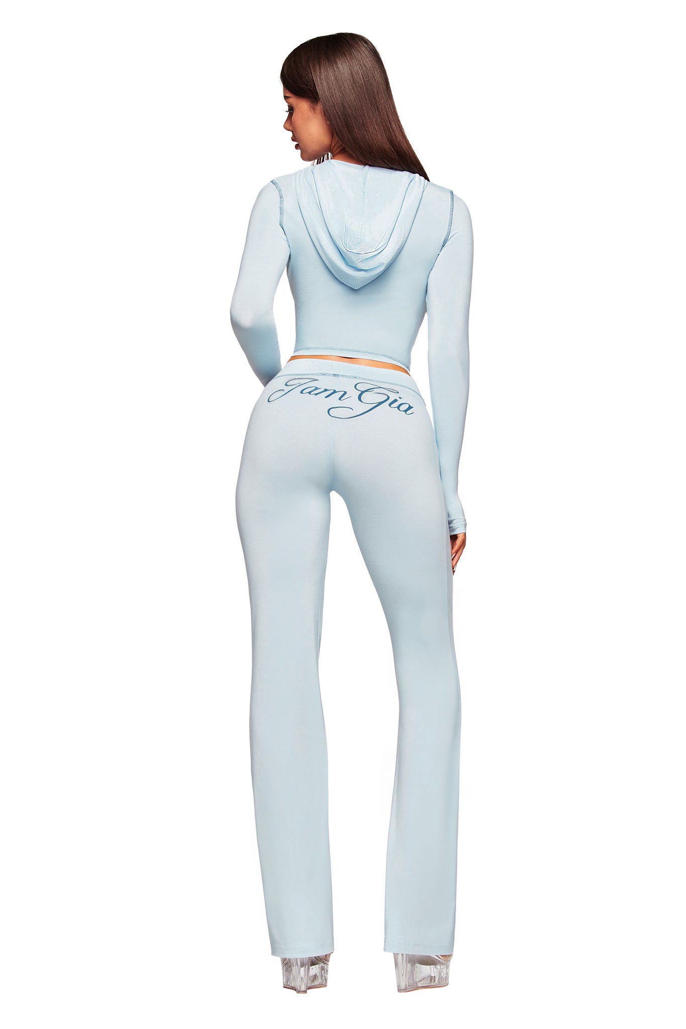 A woman with long brown hair, facing away, wears I.AM.GIA’s BLARE TRACKPANT in baby blue with flared legs and “I.AM.GIA” on the back, paired with a light blue hooded crop top and clear platform heels, set against a white background.