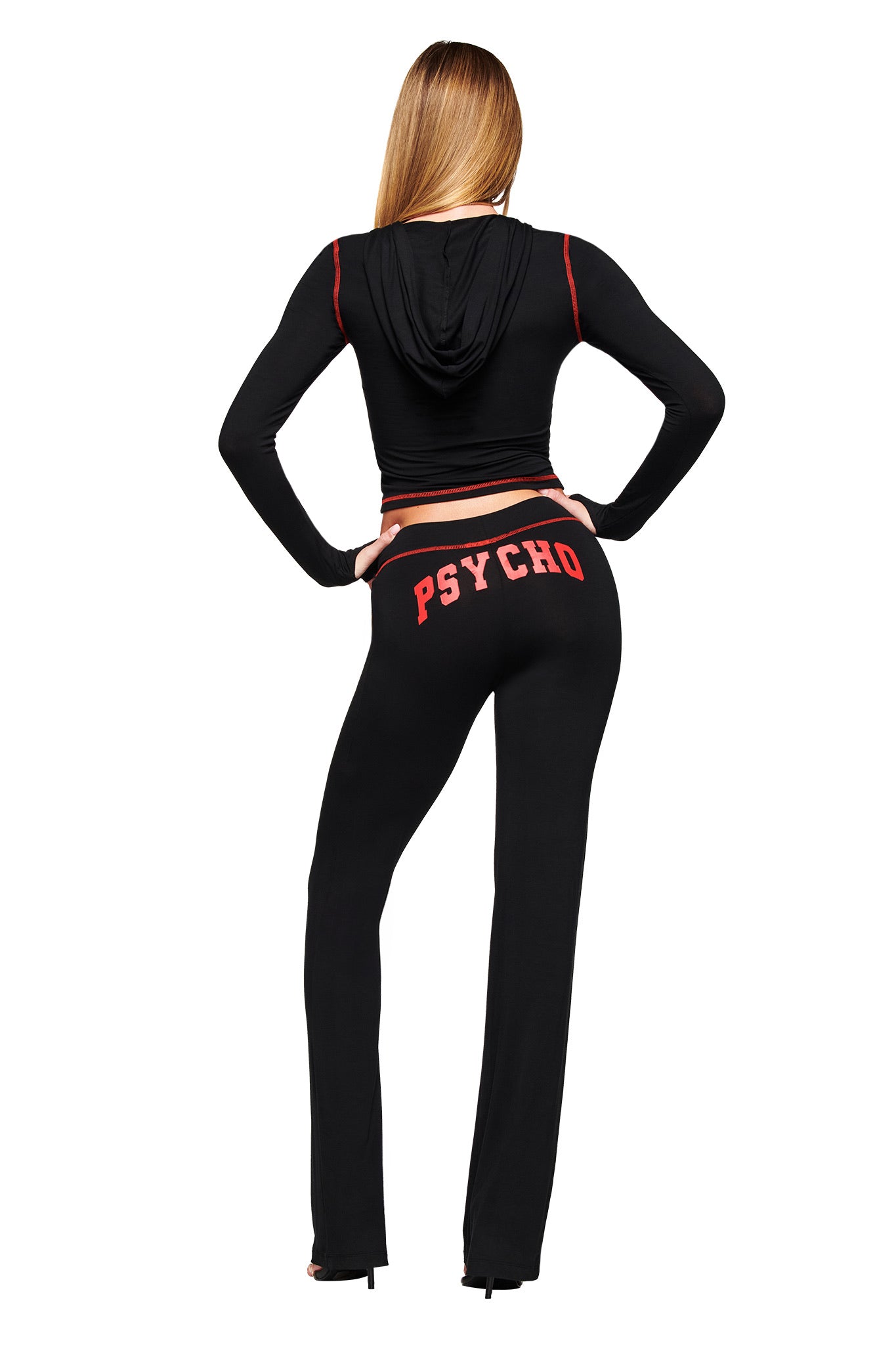 A woman with long blonde hair, hands on hips, faces away wearing I.AM.GIA's BLARE TRACKPANT - BLACK PSYCHO, featuring a bold red PSYCHO logo on the back, paired with a black cropped hoodie.