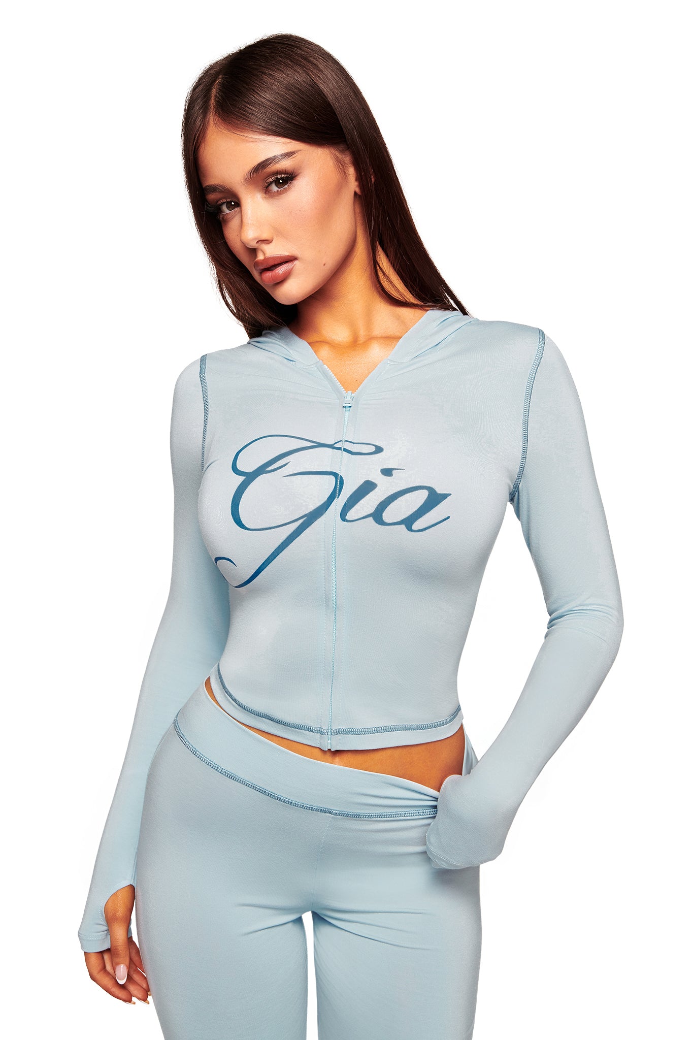 A woman with long brown hair wears the I.AM.GIA BLARE HOODIE in baby blue, featuring "Gia" on the front, and matching high-waisted leggings, standing against a white background.