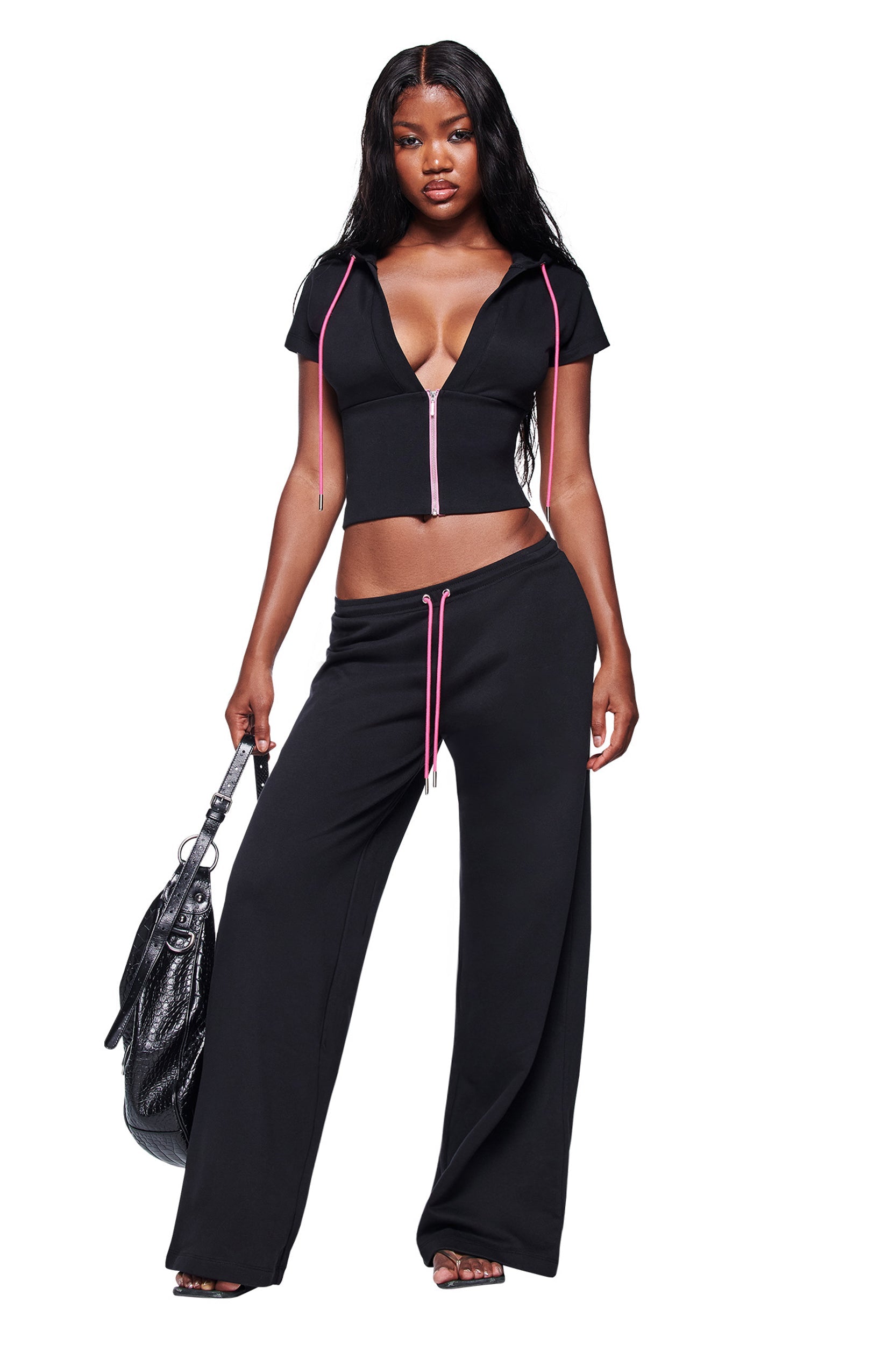 A woman stands confidently in the I.AM.GIA ARLO TRACK JACKET - BLACK with a deep V-neck and wide-leg pants featuring pink drawstrings. She holds a black handbag, her long wavy hair flowing against a white background.