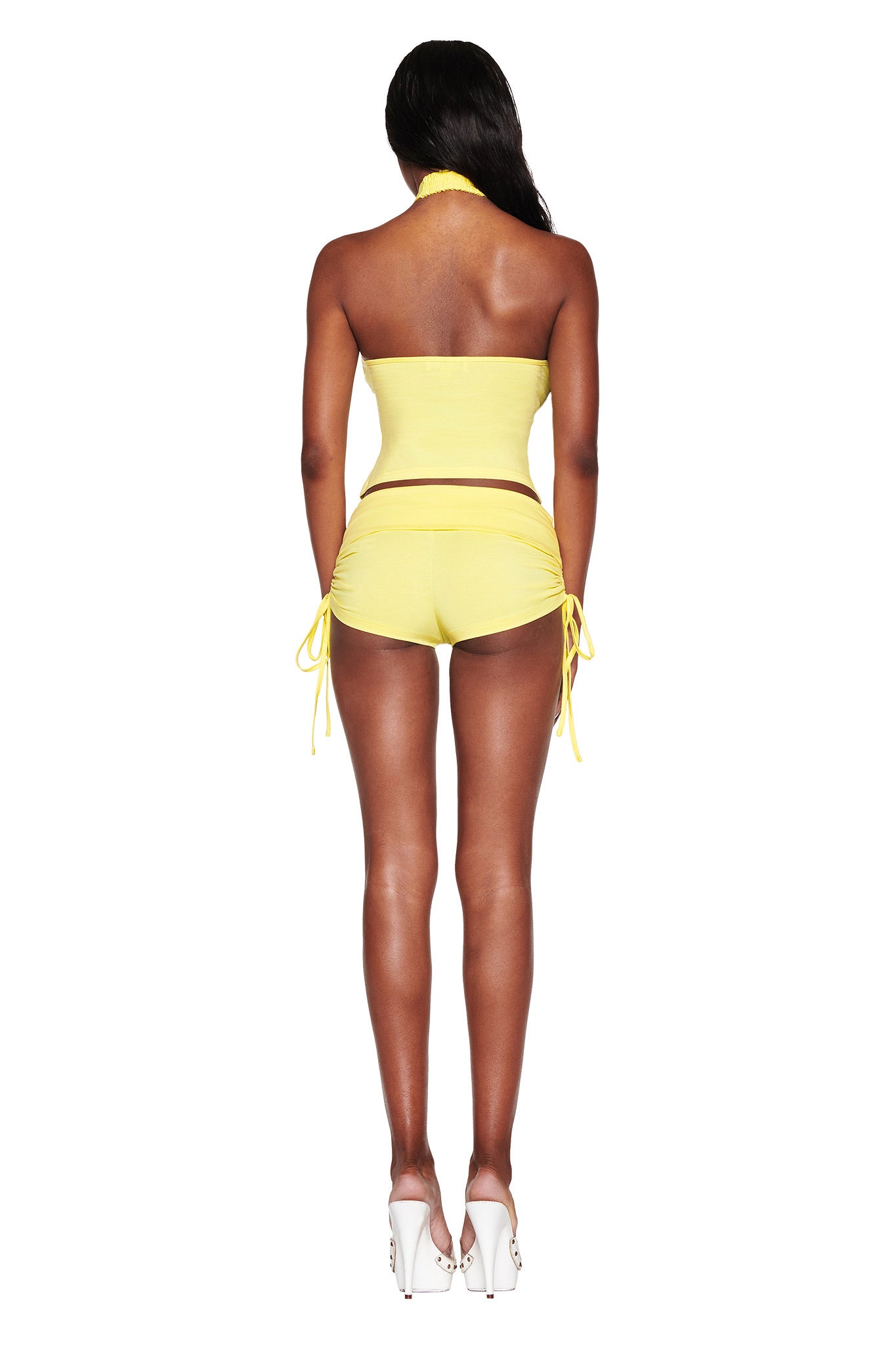 A person models the I.AM.GIA ROWAN TOP in baby yellow paired with matching shorts that have drawstring sides for a bodycon fit. They wear white high heels, and long black hair flows down their back as they face away from the camera.