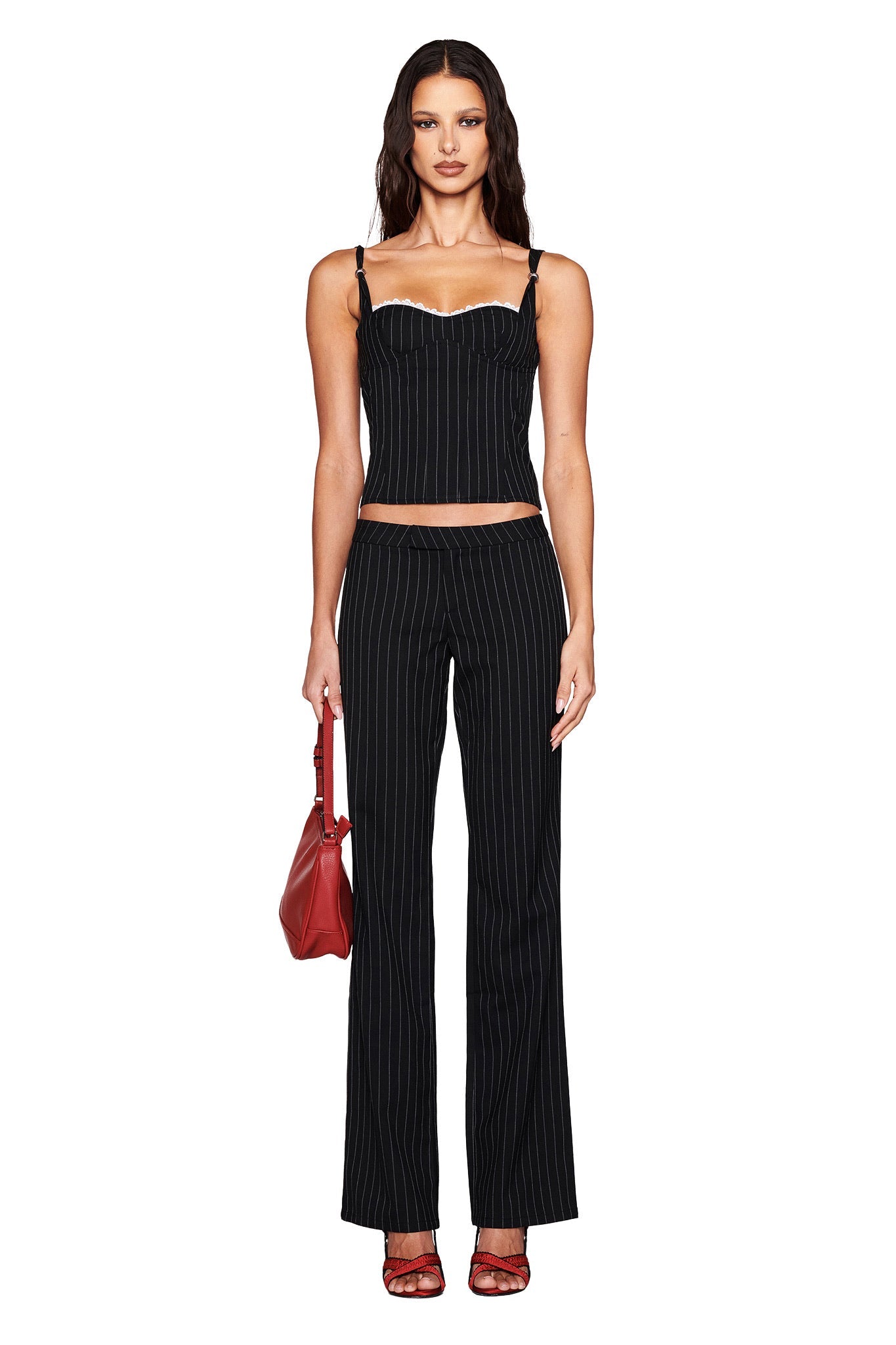 A person with long hair wears an I.AM.GIA black pinstripe corset top and MARRION PANT - BLACK straight-leg, low-rise pants while holding a red handbag against a white background, paired with red strappy heels.