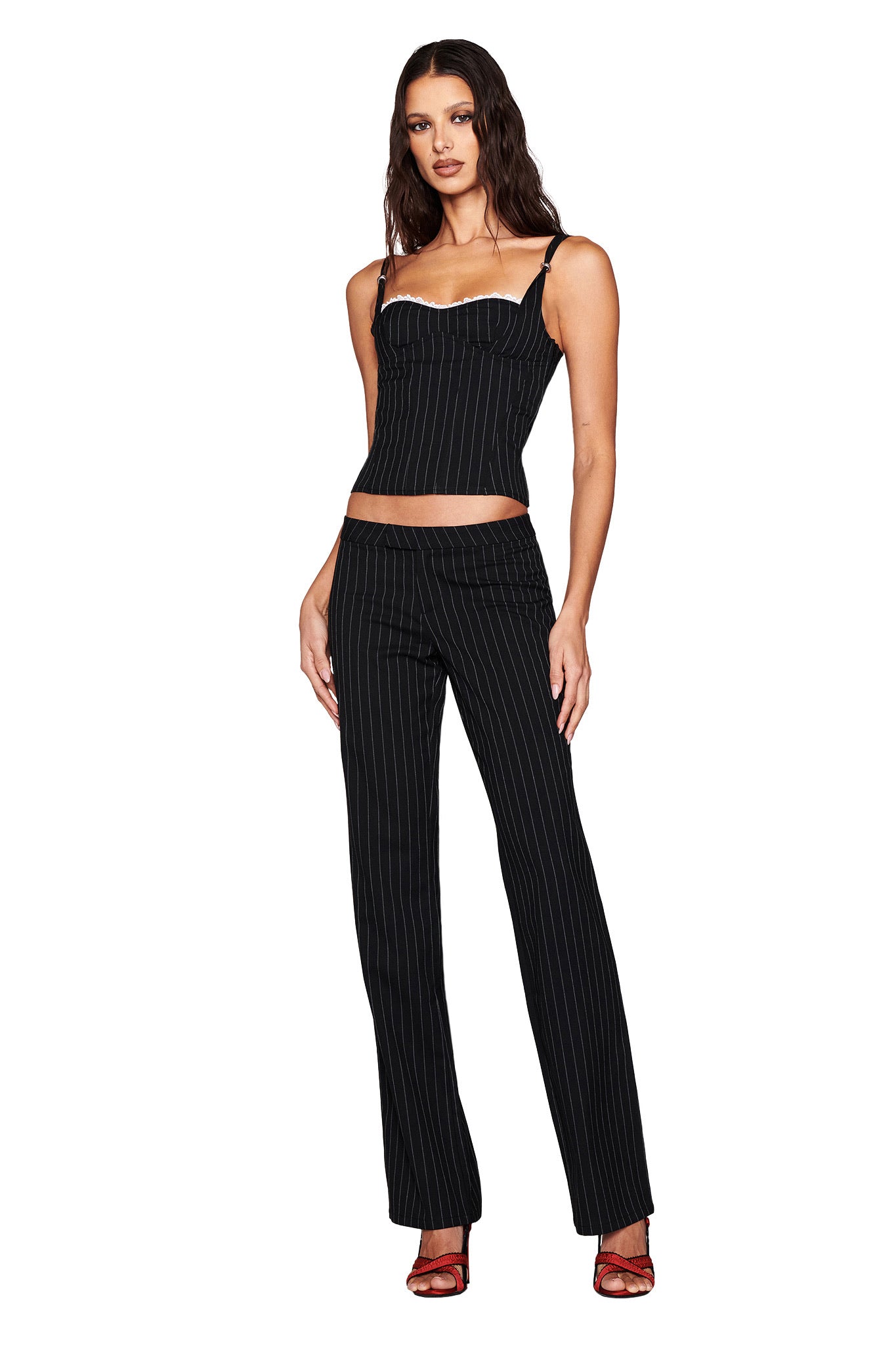 A woman wearing the black MARRION CORSET by I.AM.GIA stands against a white background. Her pinstripe outfit with a fitted under-bust top and matching pants complements her loose waves, while red strappy heels add a vibrant touch to her look.