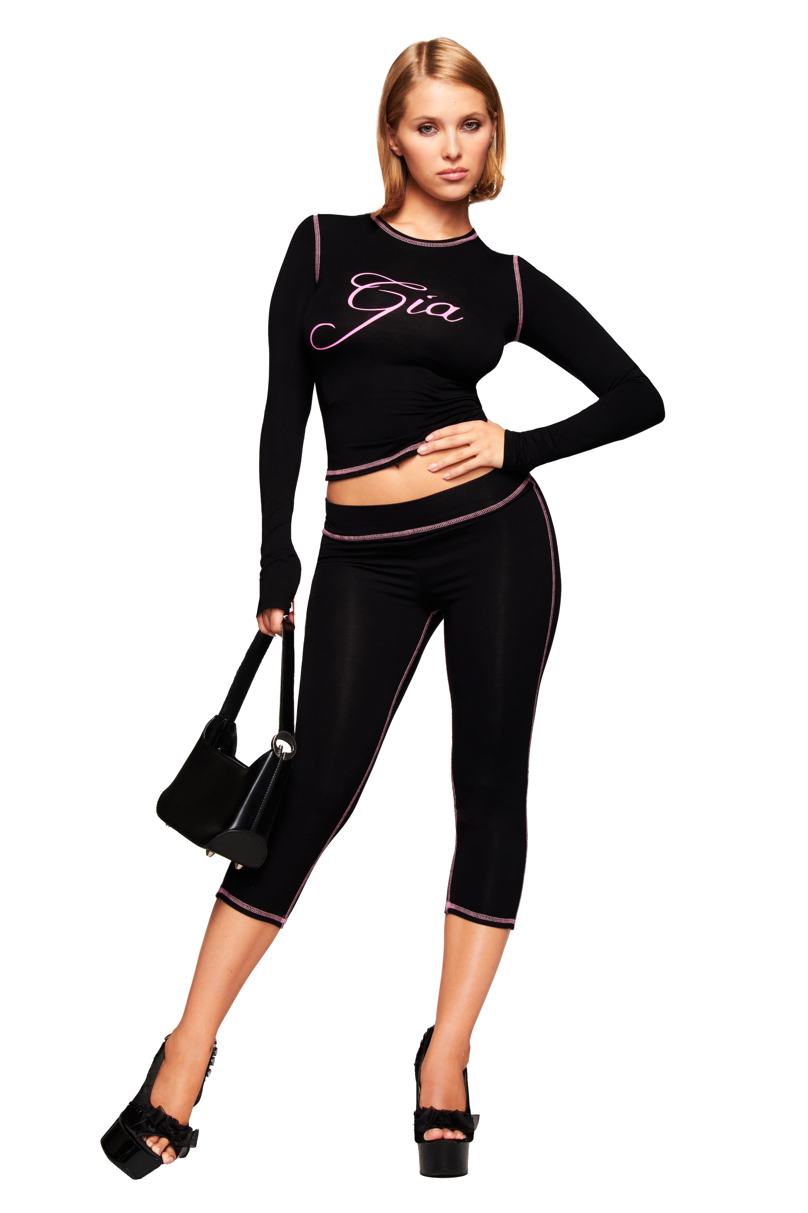 A woman poses in the I.AM.GIA BLARE CAPRI - BLACK pants and a fitted black crop top with the I.AM.GIA logo, styled with a black handbag and black platform sandals against a plain white background.