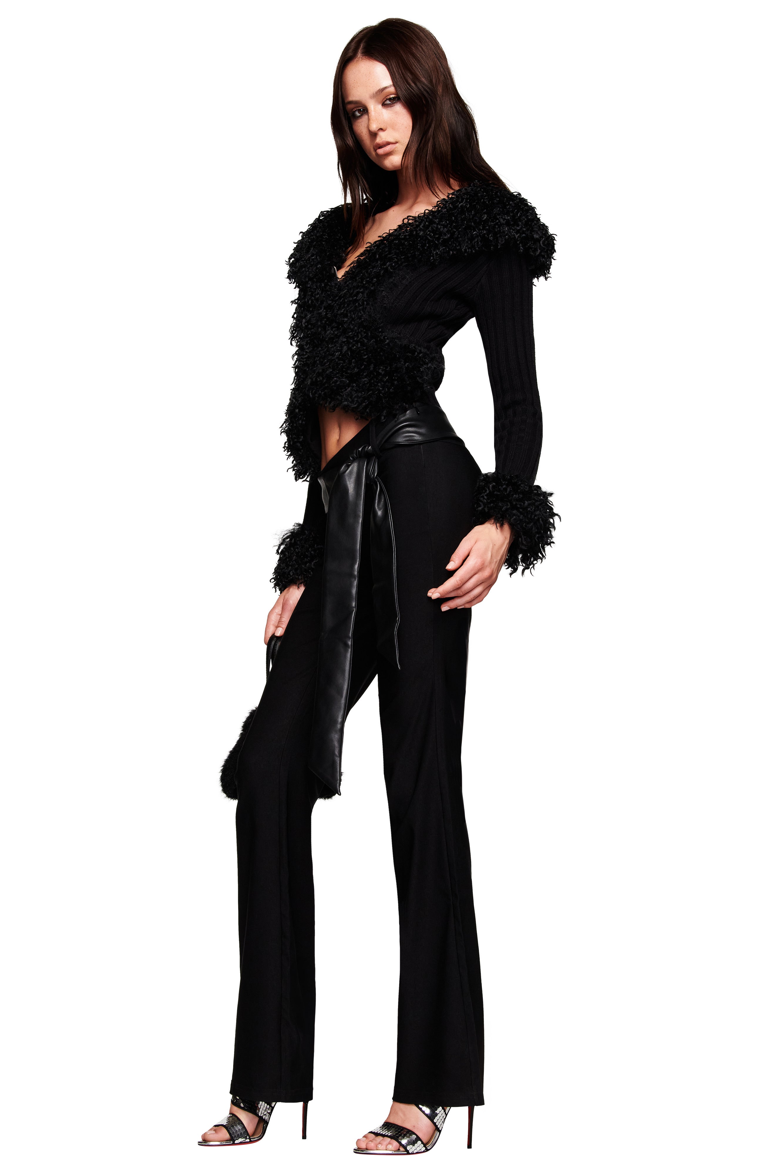 A woman models the I.AM.GIA CRESSIDA JACKET - BLACK with faux fur collar and cuffs, a black satin-like waist belt, paired with black pants and high-heeled sandals, set against a white background.