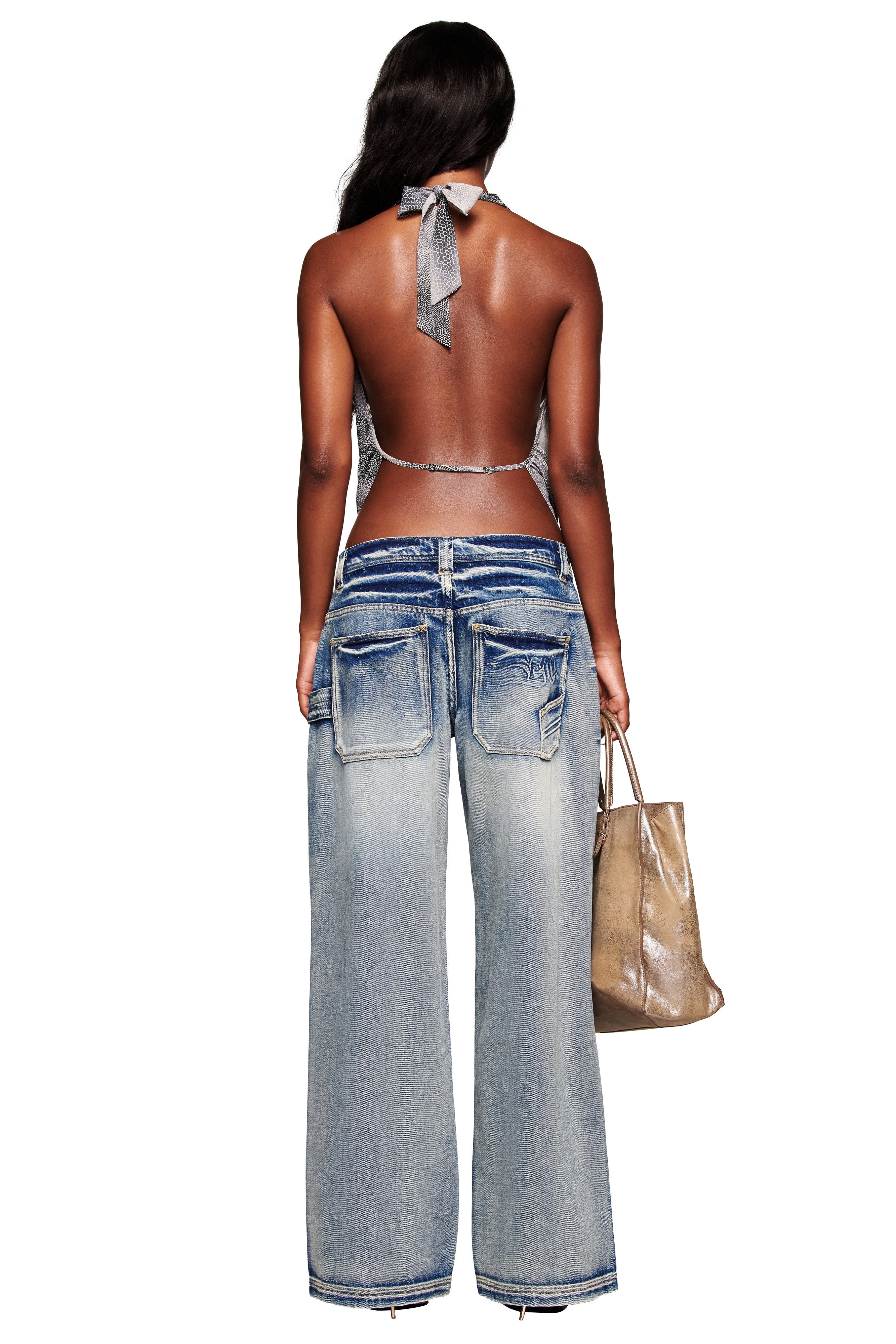 A woman with long, wavy hair stands facing away against a white background, wearing I.AM.GIA’s PALISADE JEAN - BLUE—loose, faded wide leg jeans with large back pockets—a halter top, and holding a metallic tote in her left hand.