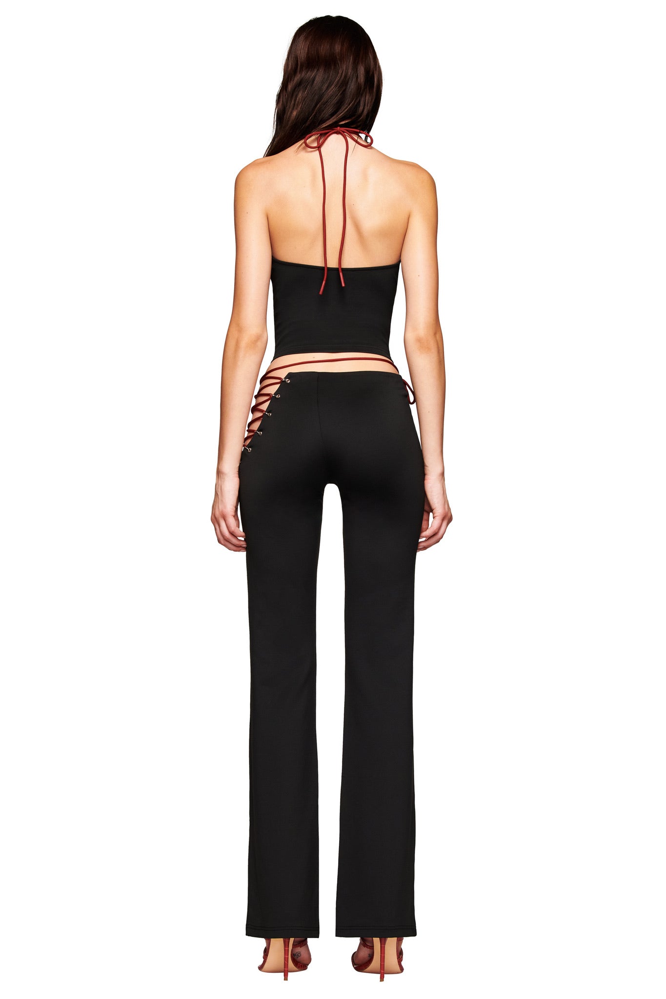 A woman stands with her back to the camera in a black halter top and I.AM.GIA ONYX PANT - BLACK, featuring low-rise fit, eyelet fabric, cutout hips, and open sides. She wears high-heeled shoes against a white background.