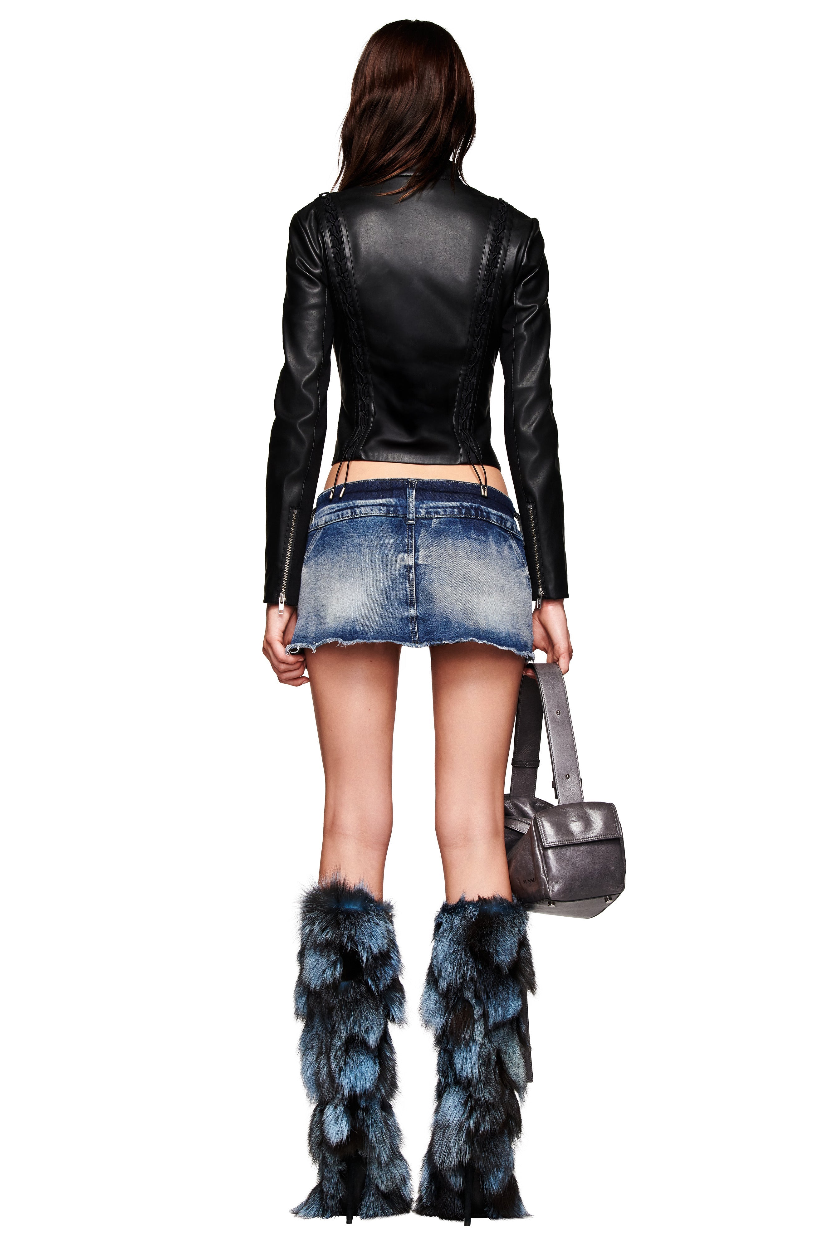 A woman is seen from behind wearing the I.AM.GIA KENDRIX JACKET in black, a short denim skirt, black underwear, tall blue and black faux fur boots, and holding a silver handbag.