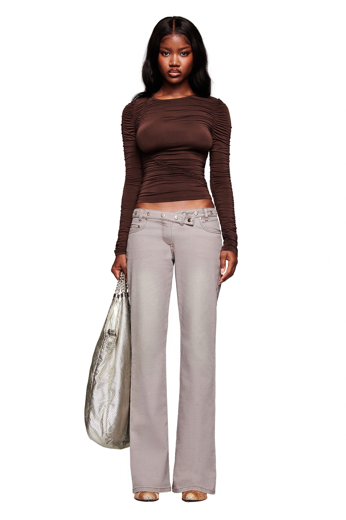 A woman poses against a white background wearing the I.AM.GIA GREER PANT in grey, styled with a fitted long-sleeve brown top, a large metallic silver bag, and light-colored open-toe shoes.