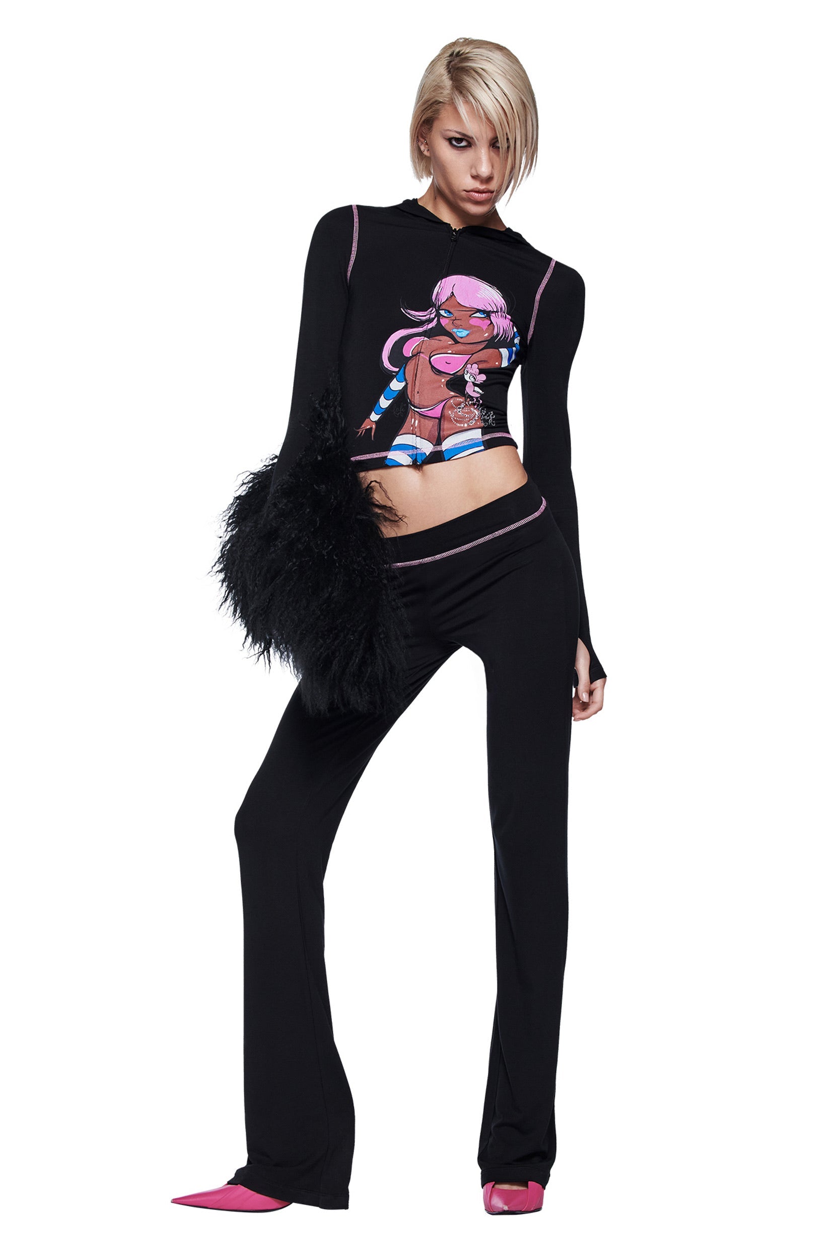 A person with short blonde hair poses in a fitted black outfit featuring an I.AM.GIA FAFI BLARE PANT - BLACK, a cartoon graphic top, fluffy bag, and pointed pink heels against a white background.