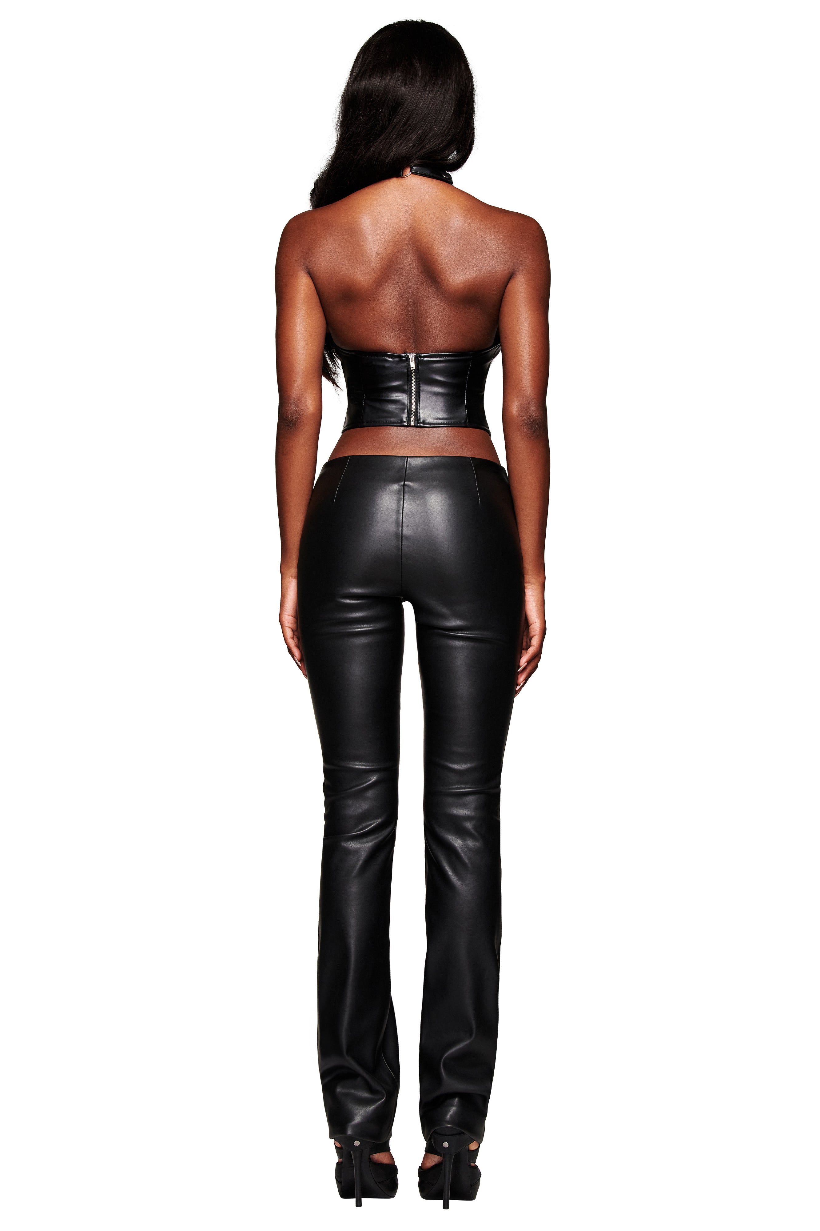 A woman faces away from the camera in I.AM.GIA’s KENDRIX PANT - BLACK, a low-rise, straight-leg faux leather moto pant, styled with a black strapless crop top and heels against a white background.