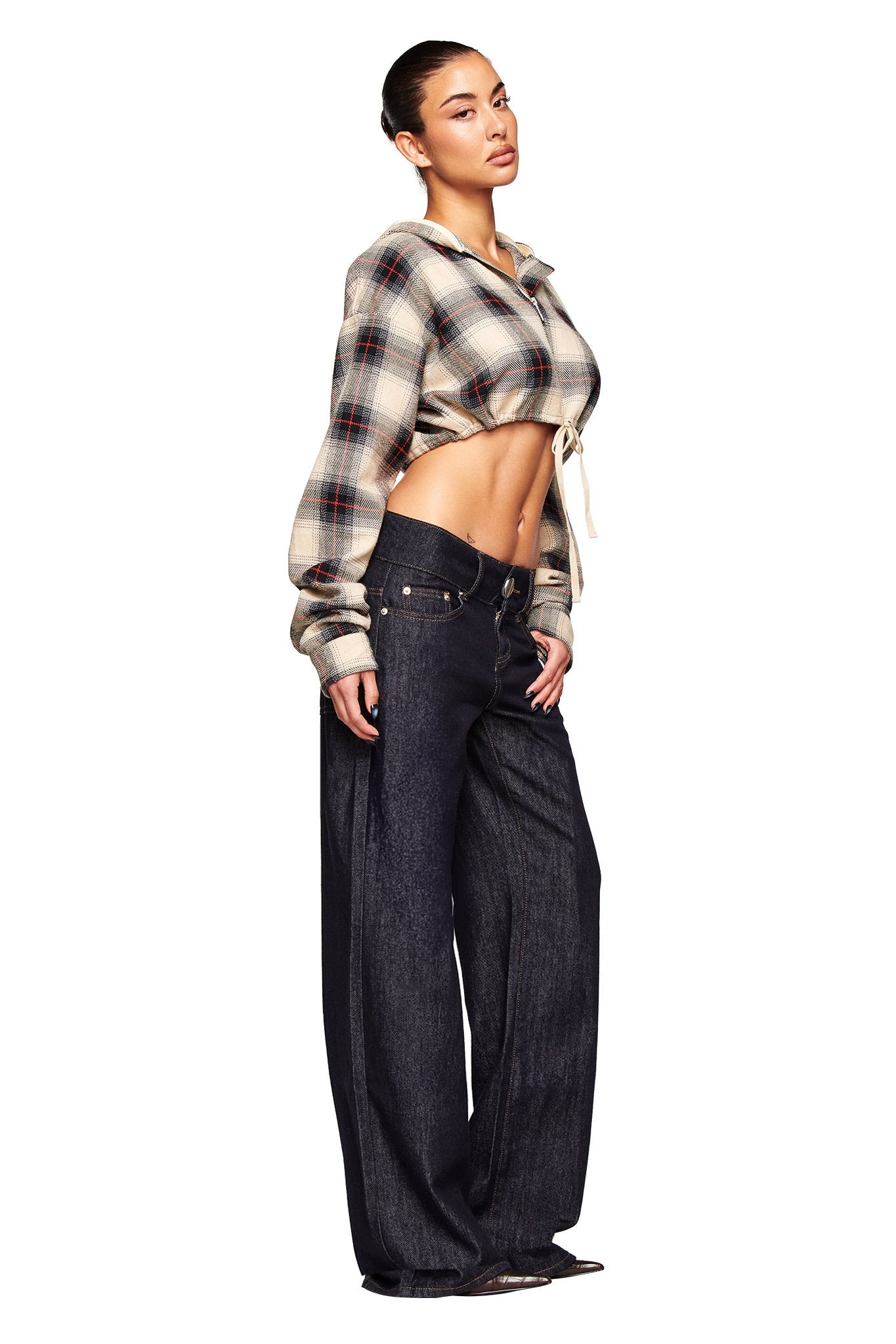 A woman poses confidently against a white background wearing I.AM.GIA’s MOXIE JEAN - INDIGO, styled with a cropped plaid hoodie that reveals her midriff. Her hair is tied back, highlighting the jeans’ low rise and wide waistband design.