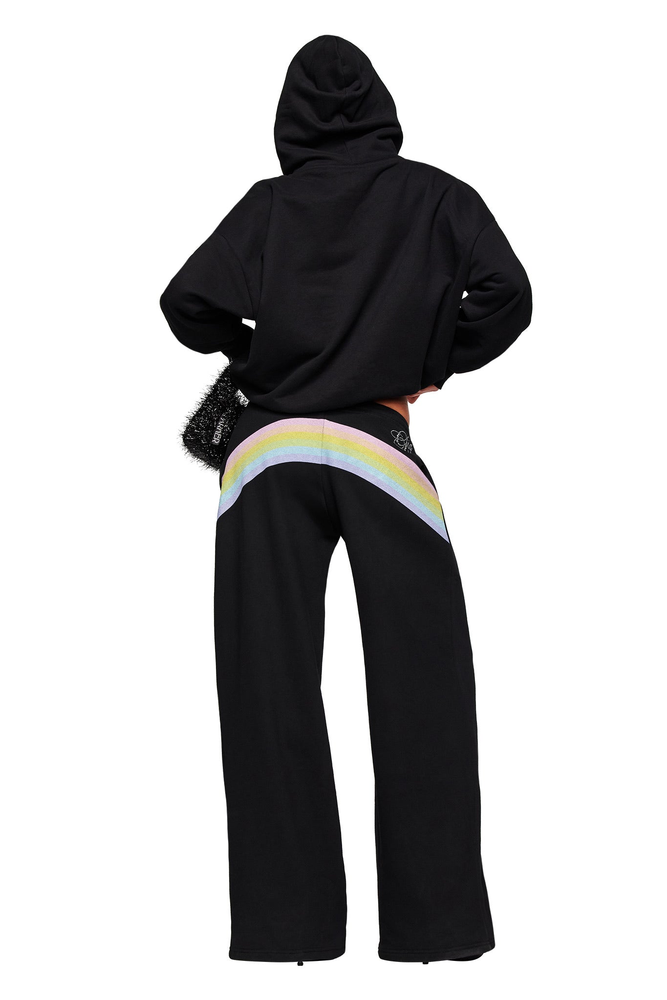 Wearing the I.AM.GIA FAFI HOODIE in black and matching pants with a pastel rainbow design, a person stands with hands on hips, back to the camera—showcasing the brand's bold streetwear aesthetic.