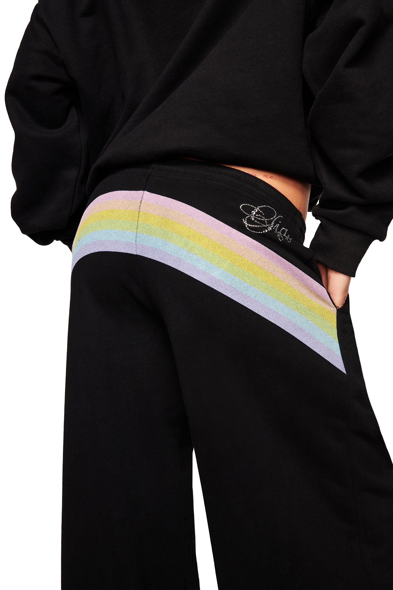 Model wears the I.AM.GIA FAFI TRACKPANT in black, featuring a pastel rainbow stripe across the back and an embroidered logo near the low-rise waist, shown from waist down and slightly turned to the side.