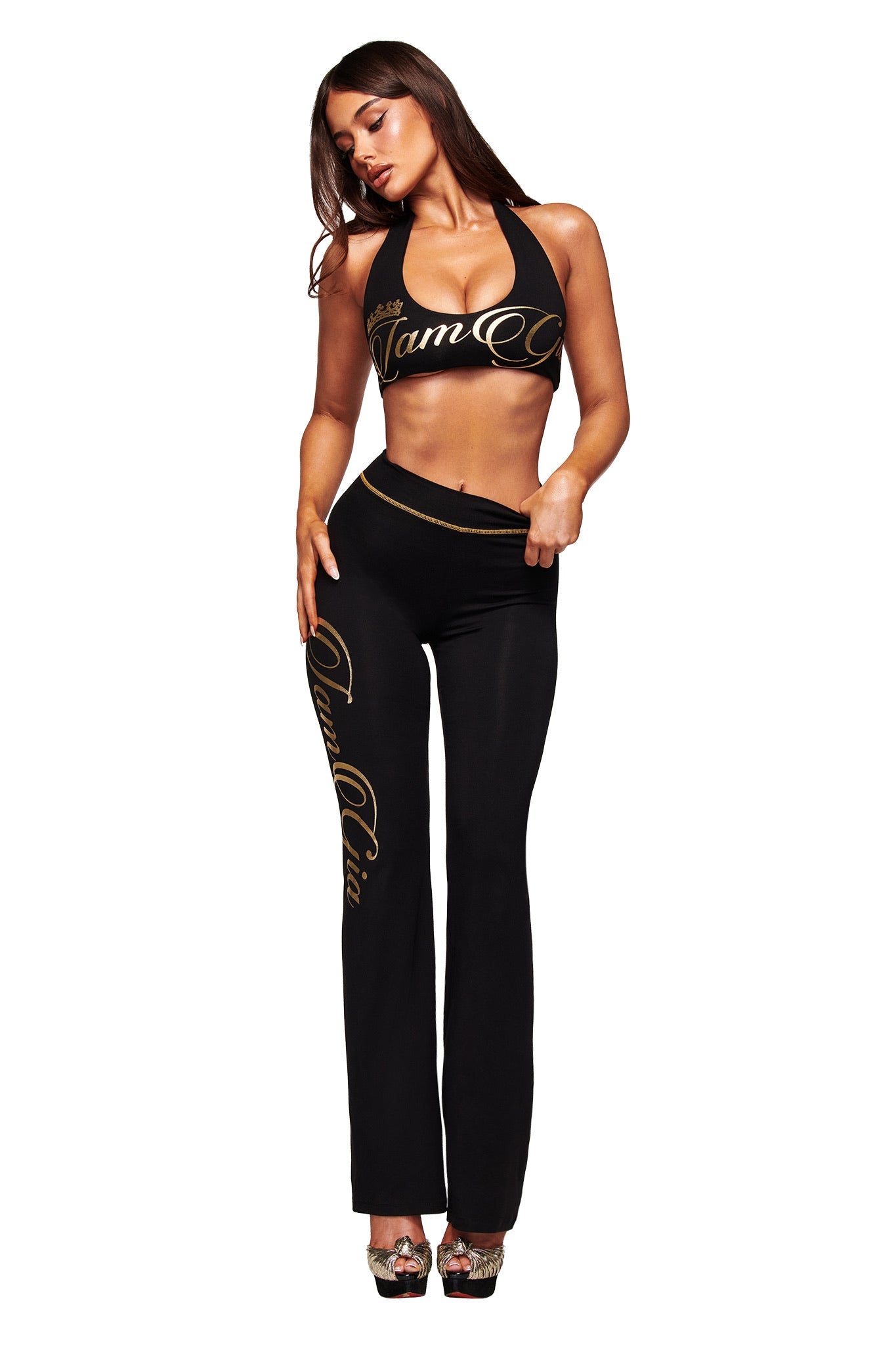 A woman models the I.AM.GIA BLARE FORM HALTER CROP TOP in black and gold, paired with matching pants and high heels, against a white background.